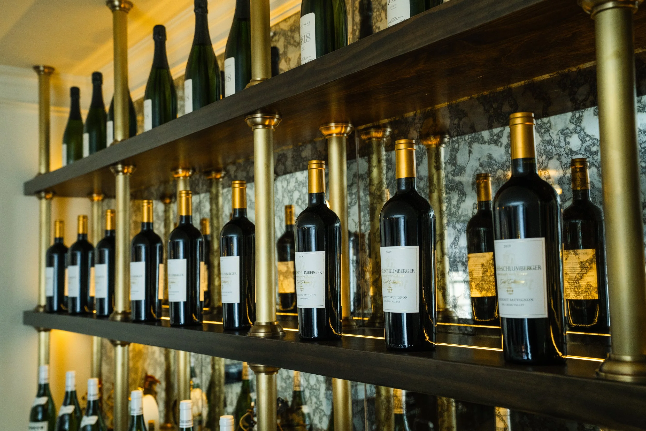 Rows of wine bottles on a shelf with gold accents in a dimly lit setting.