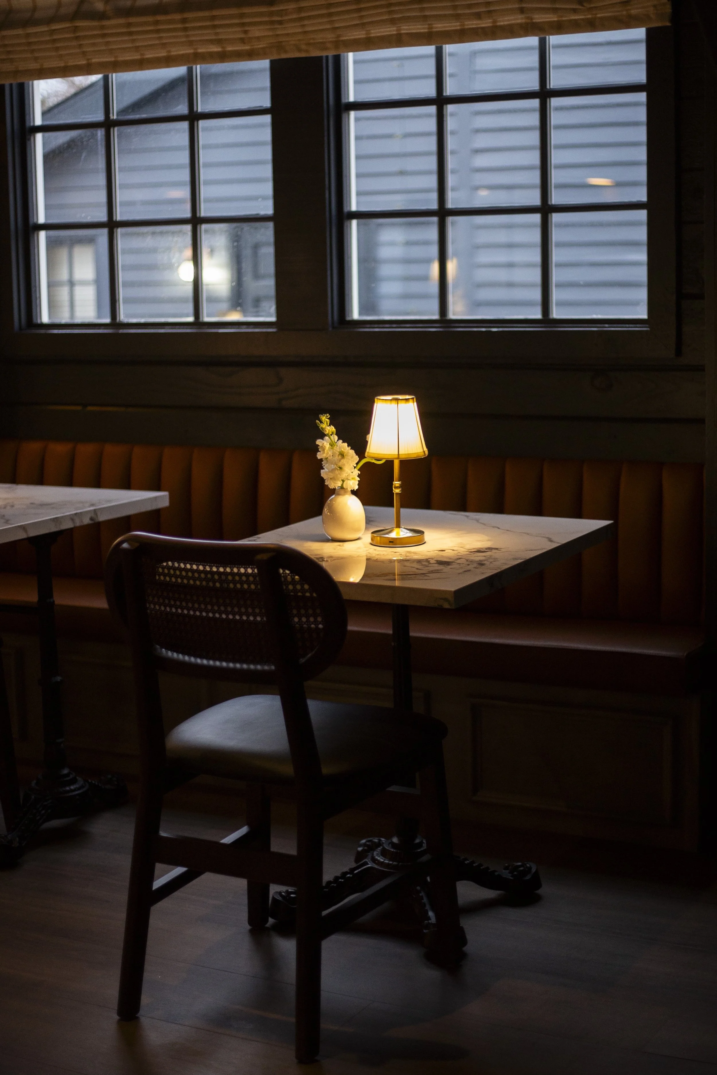 Cozy café interior with a table, chair, small table lamp, and a vase with flowers by a window.