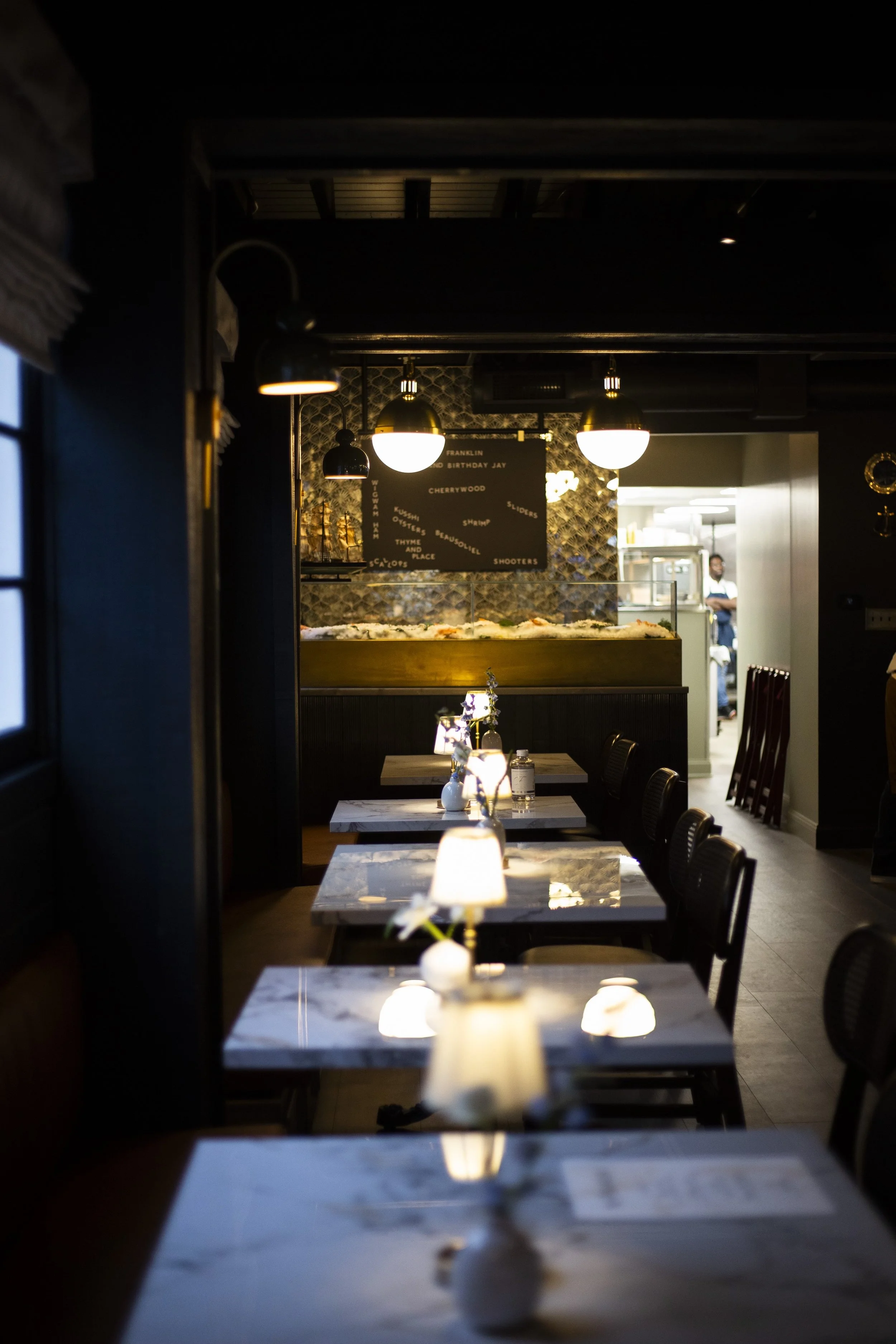 Interior of a dimly lit restaurant with marble tables, small lamps, and a view into the kitchen. Decorative lighting and wall art enhance the ambiance.