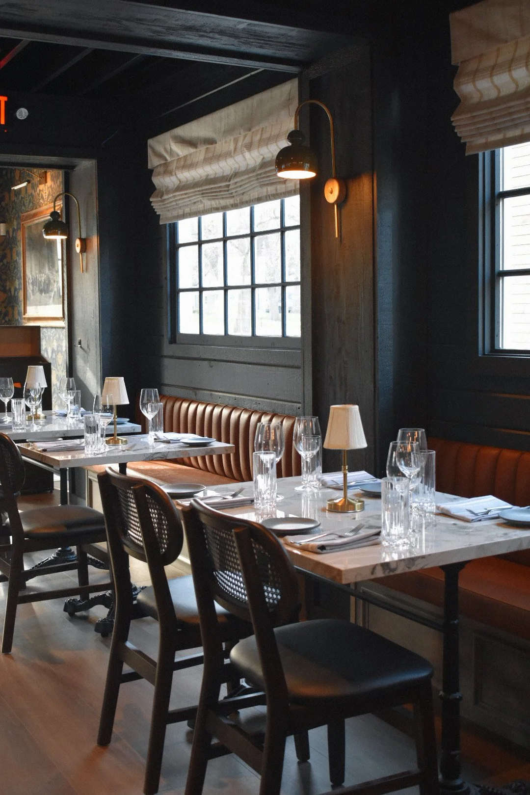 Cozy restaurant interior with wooden tables, set with glassware and napkins, leather seating, cushioned chairs, and wall lamps. Large windows provide natural light.