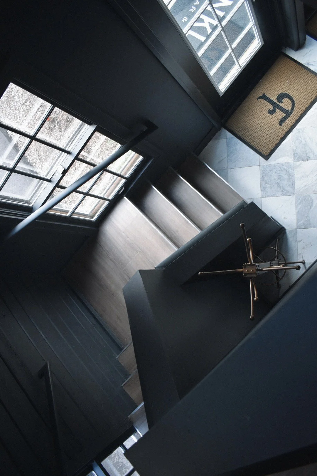 Modern interior staircase with black metal railing, wooden steps, large windows, and marble tile floor at the entryway, featuring a doormat with a decorative design.