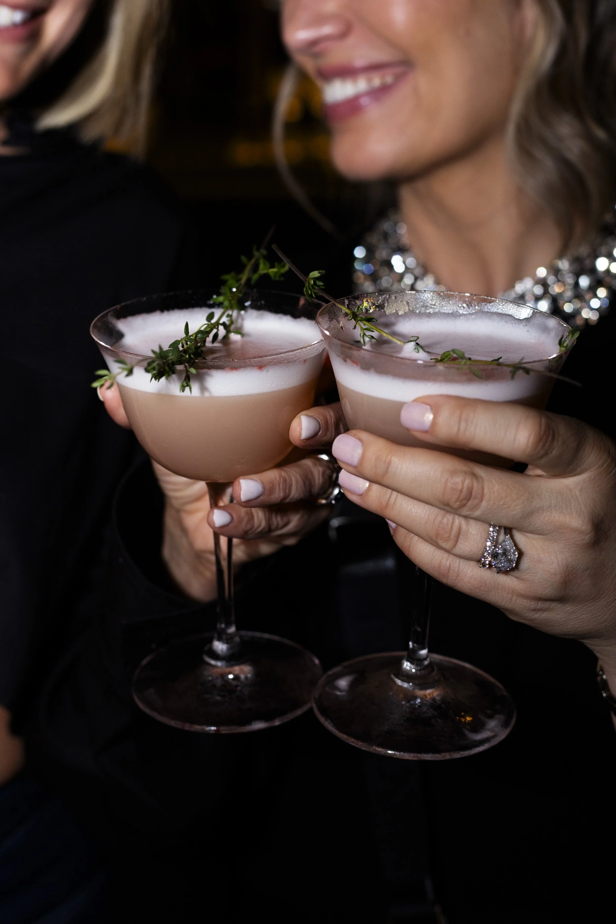 Two women holding frothy cocktails garnished with sprigs of herbs, with one woman smiling and wearing a jeweled necklace.