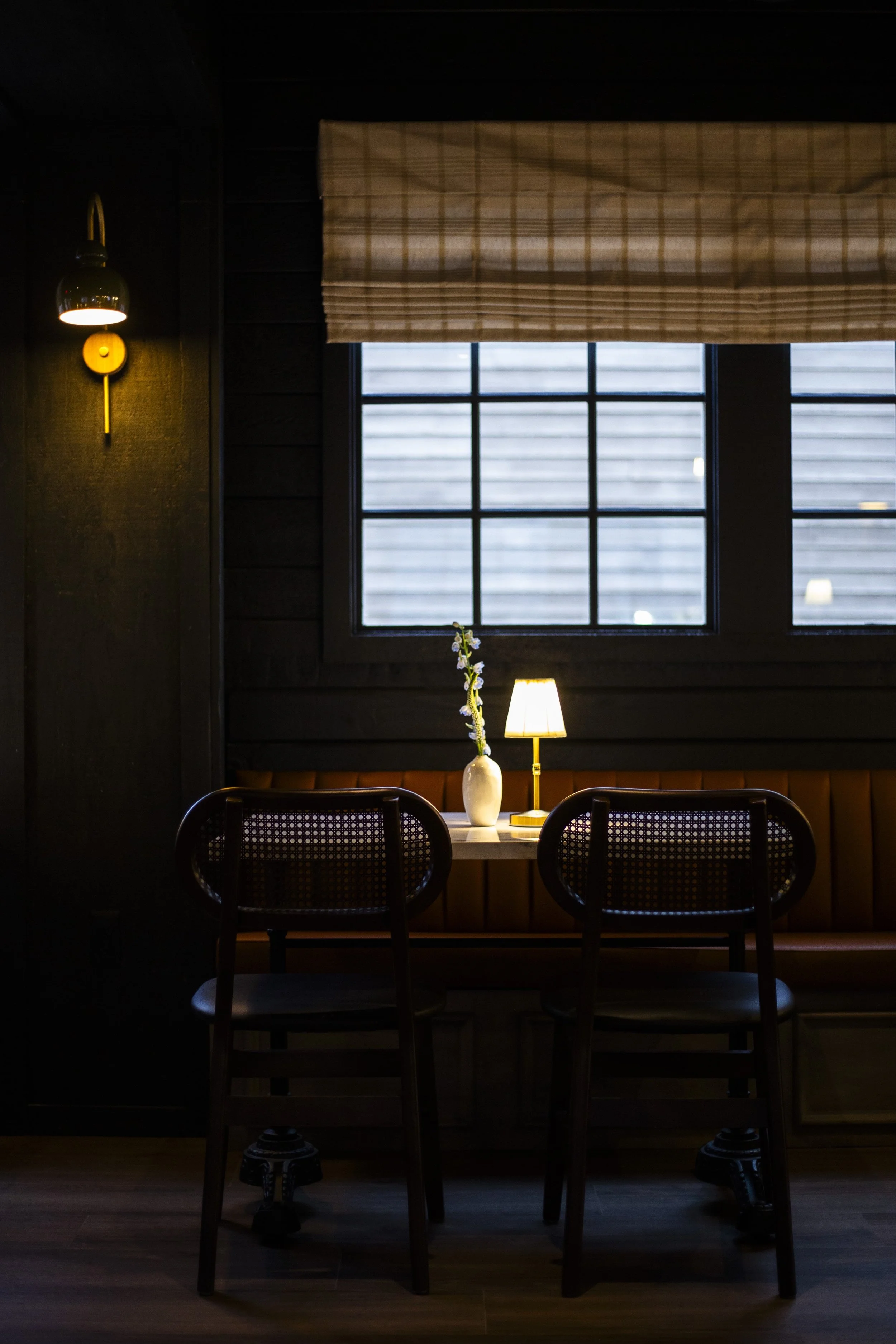 Cozy restaurant interior with wooden chairs, a small table, a table lamp, and a decorative vase on a window ledge. Warm lighting and a window with curtains create an intimate atmosphere.
