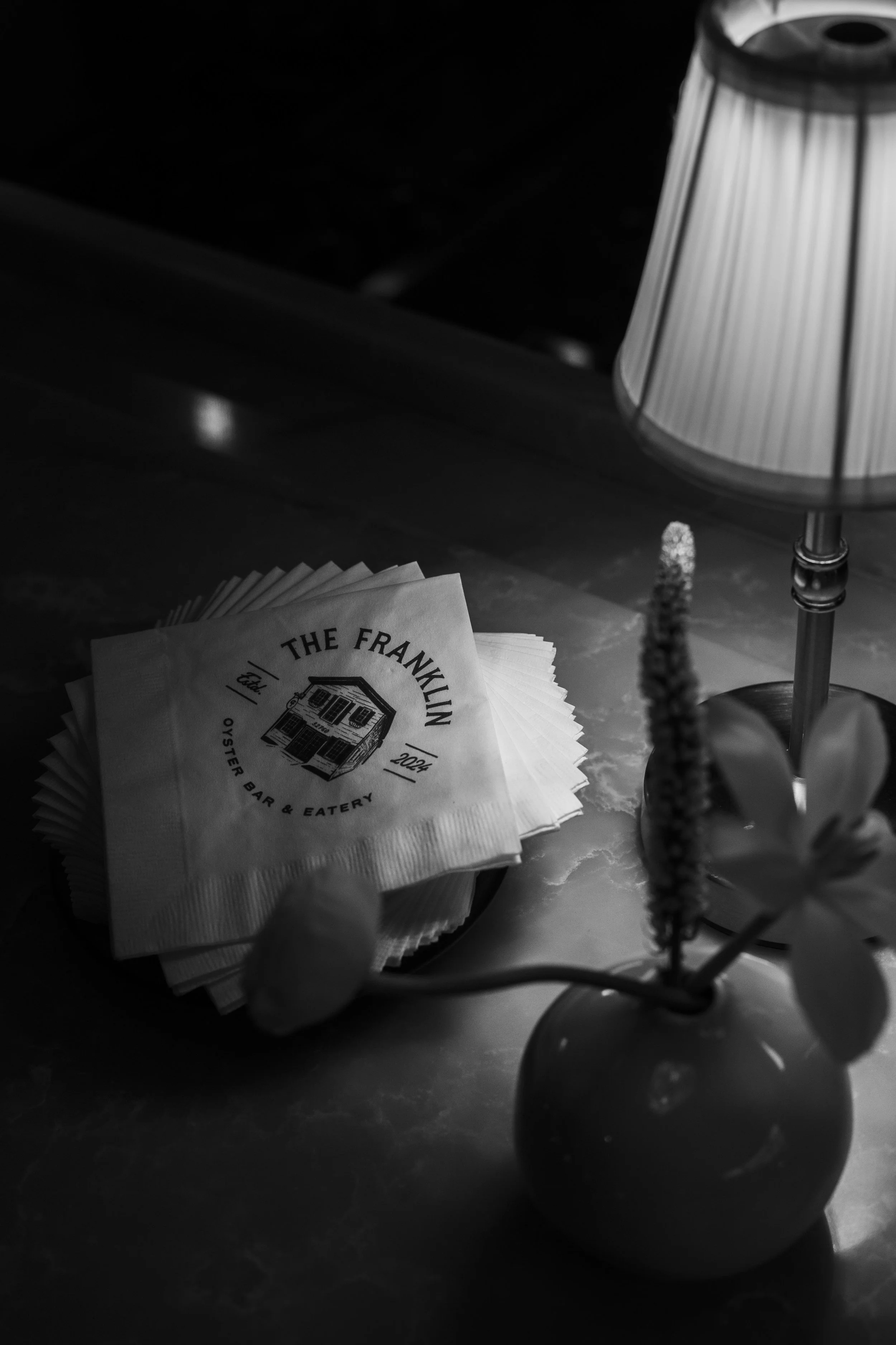 Black and white photo of a table with a stack of napkins labeled "The Franklin Oyster Bar & Eatery," a small lamp with a pleated shade, and a round vase holding a flower.