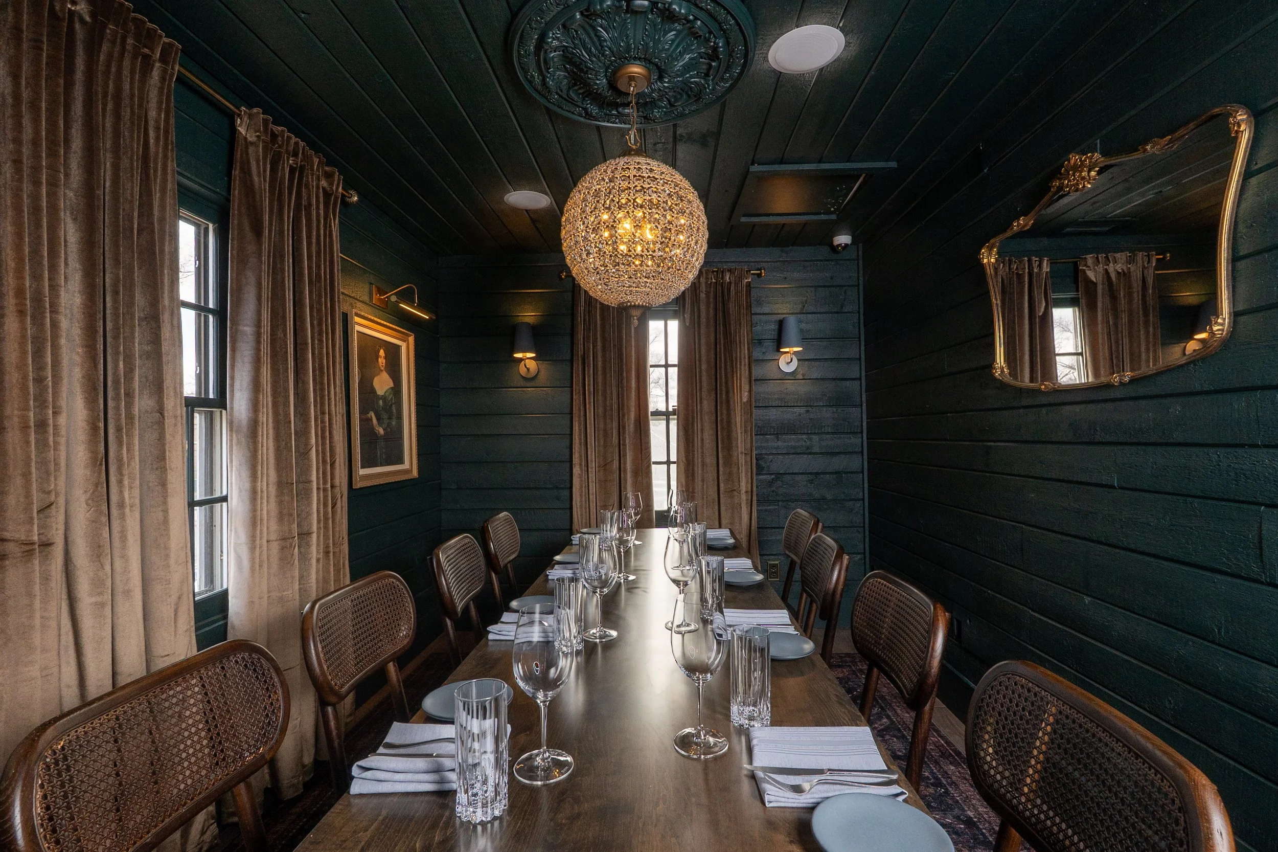 Elegant dining room with a long wooden table set for a meal, surrounded by cane chairs. A chandelier hangs above, and velvet curtains cover the windows. Dark green walls feature a painting and a decorative mirror.
