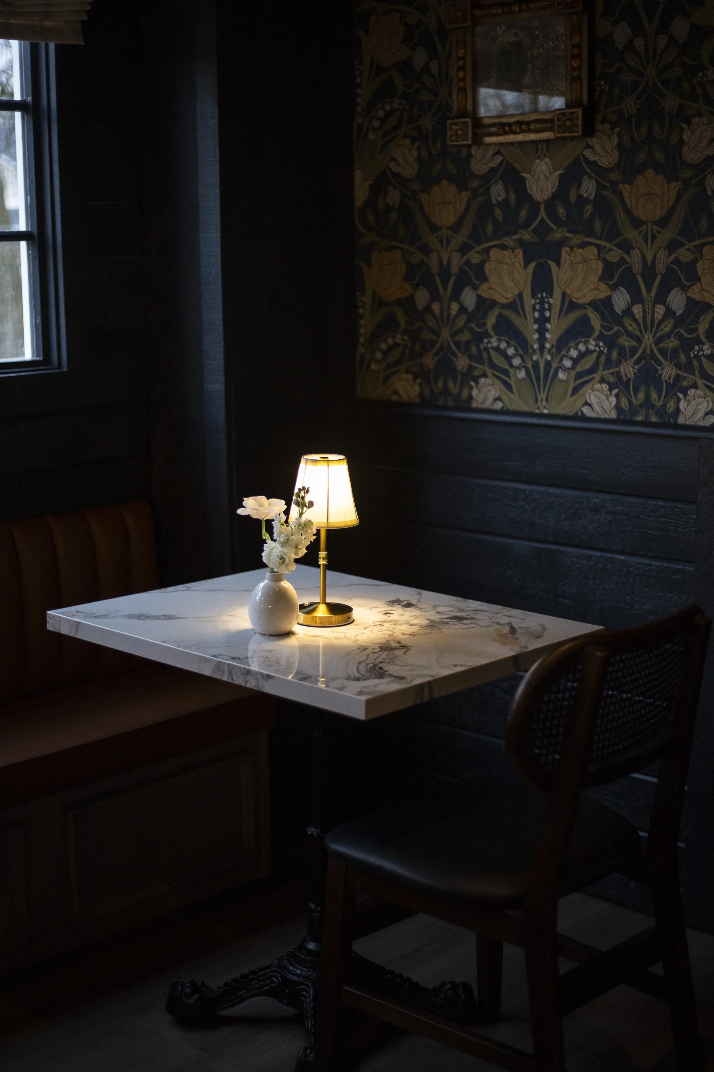 Cozy cafe corner with a marble table, a small lamp, a vase with white flowers, and dark, floral wallpaper.