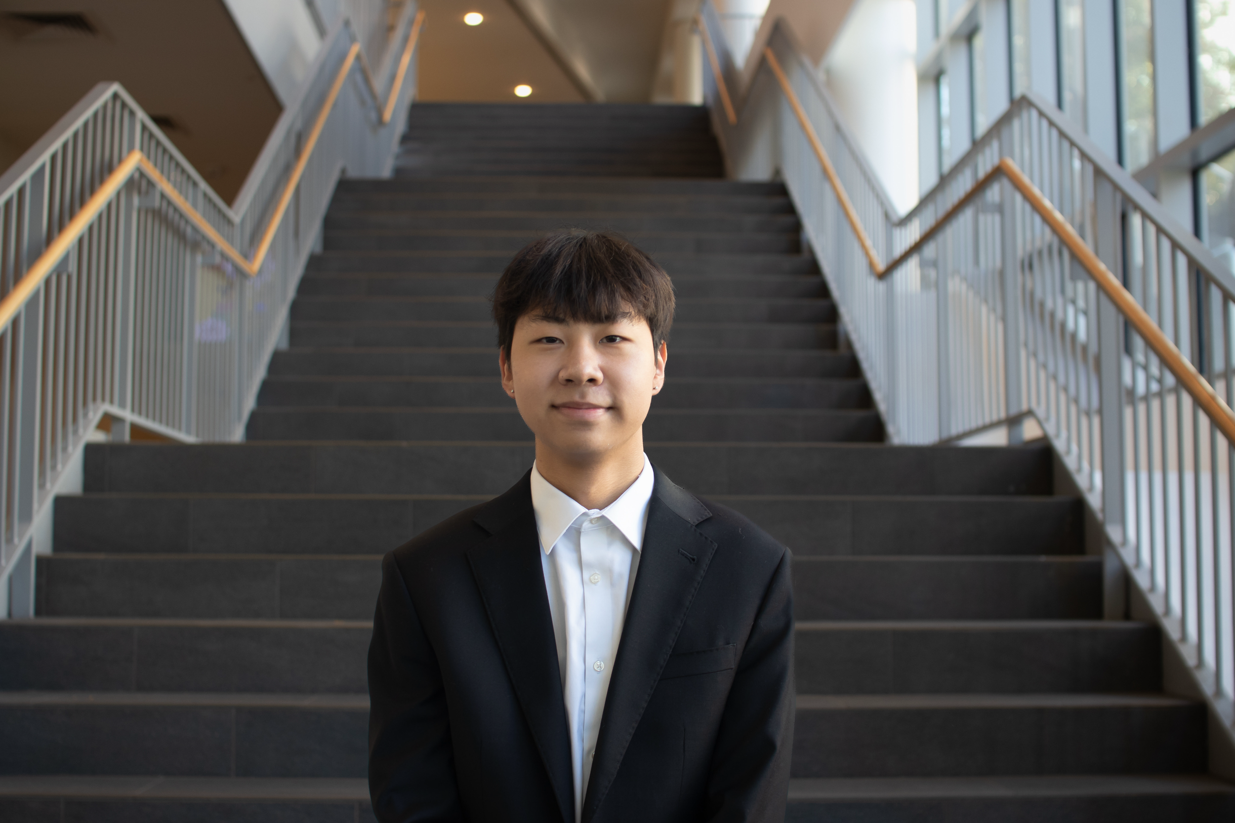 Young man in a black suit standing in front of a staircase in a well-lit building.