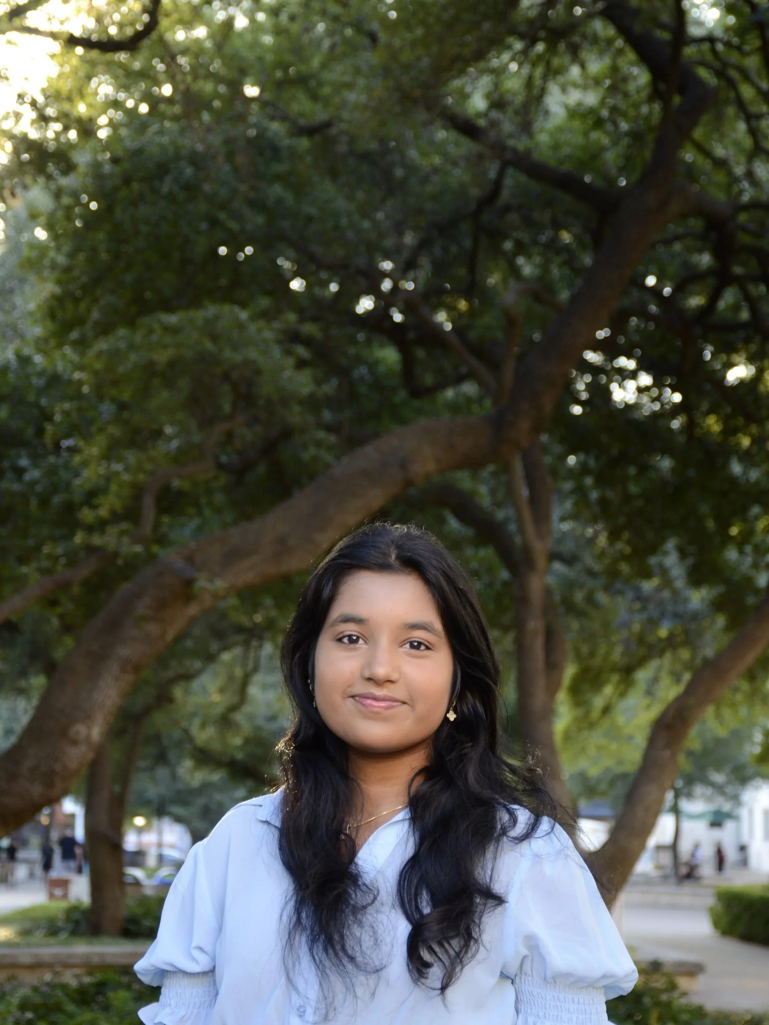 A young girl with long black hair wearing a white blouse standing outdoors with green trees and park benches in the background.