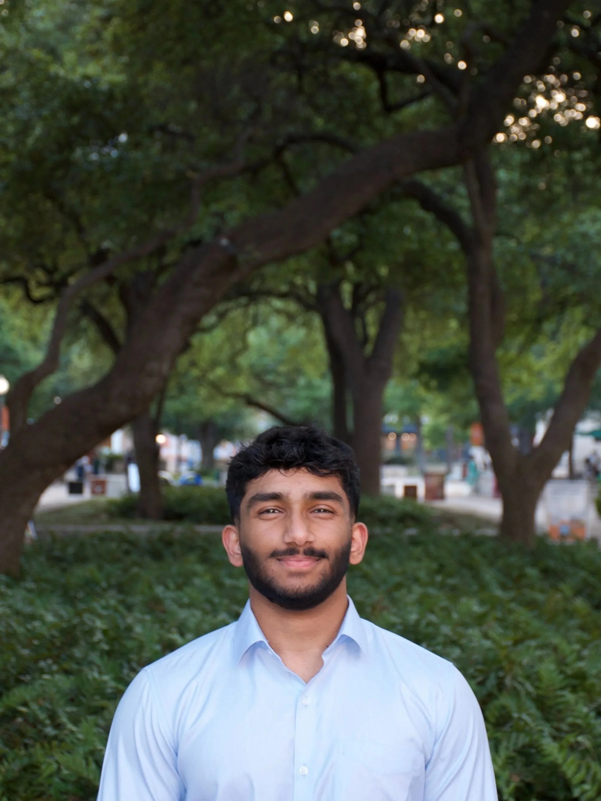 A young man with dark curly hair, a beard, and wearing a light blue button-up shirt stands outdoors in a park with lush green trees and bushes in the background.