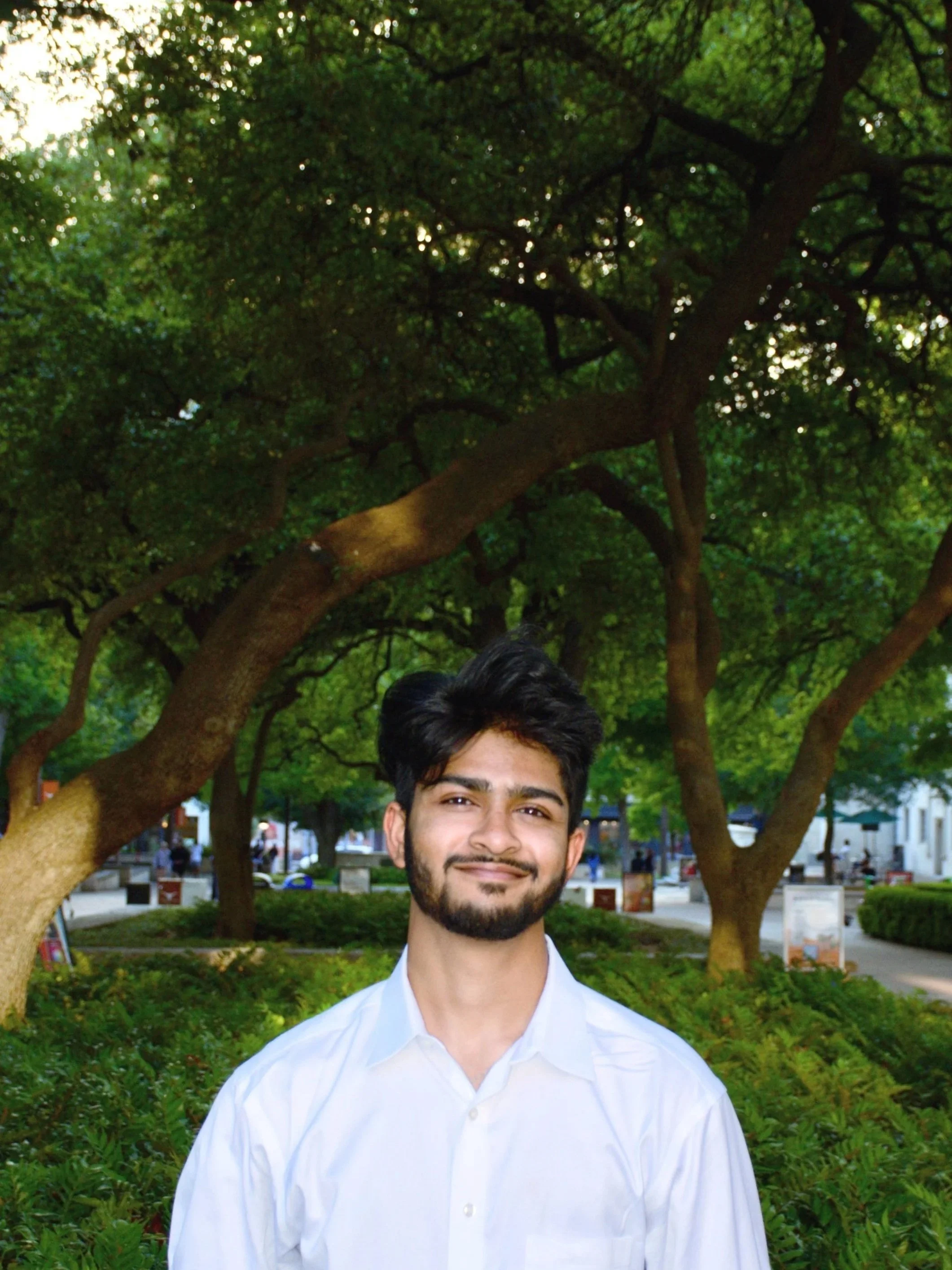 A young man with dark hair and a beard, dressed in a white shirt, smiling outdoors in a park with green trees and bushes, and a walkway in the background.