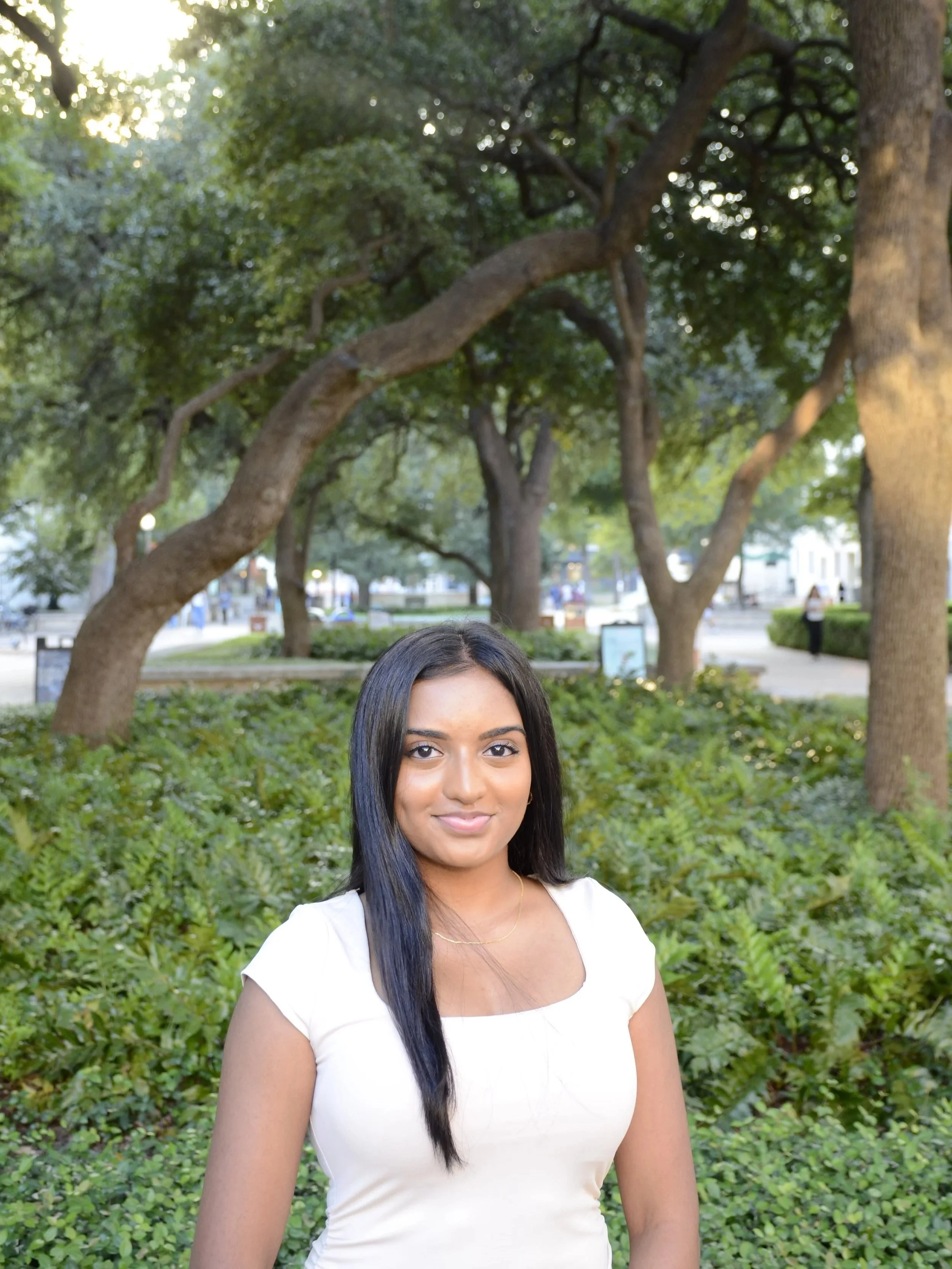 A young woman with long black hair, wearing a white top, standing outdoors in a park with trees and greenery in the background.