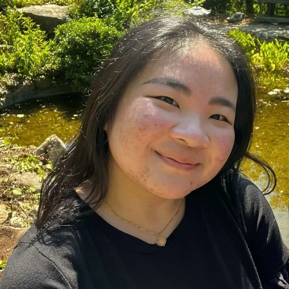 Young woman with dark hair smiling outdoors near a pond, surrounded by greenery, wearing a black top and a gold necklace.