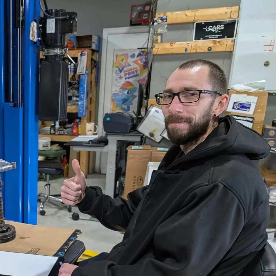 Black Subaru with bronze alloy wheels parked in a lot at a Subaru repair shop, with flags and other vehicles in the background.