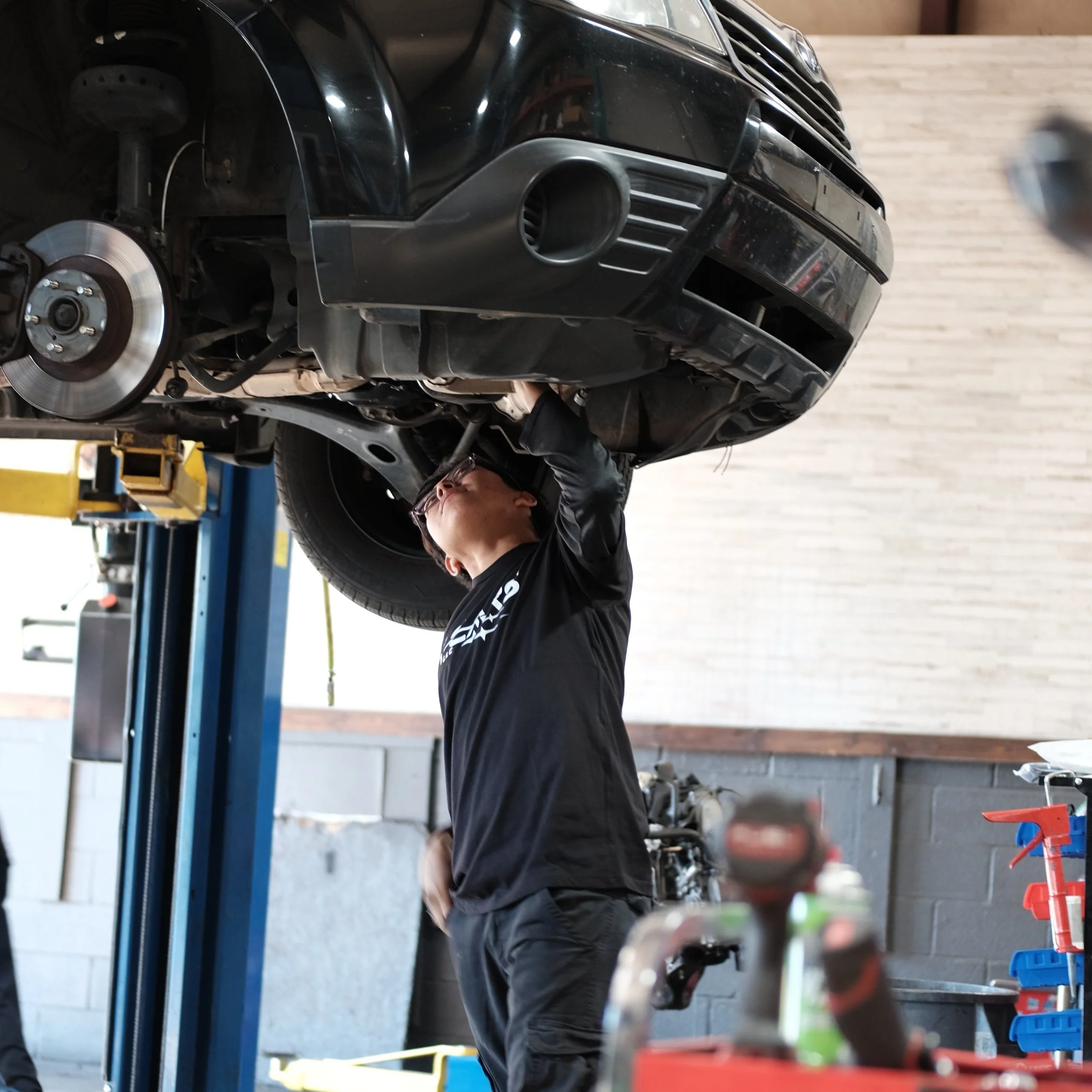 A mechanic working underneath a black car that is lifted on a hydraulic lift in an auto repair shop.