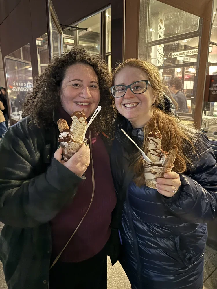 Two women smiling and holding large cups of ice cream sundaes topped with whipped cream and chocolate, in an indoor setting with city lights outside.