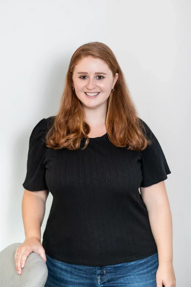 A young woman with long red hair, wearing a black short-sleeved top and blue jeans, smiling and standing against a plain white background.