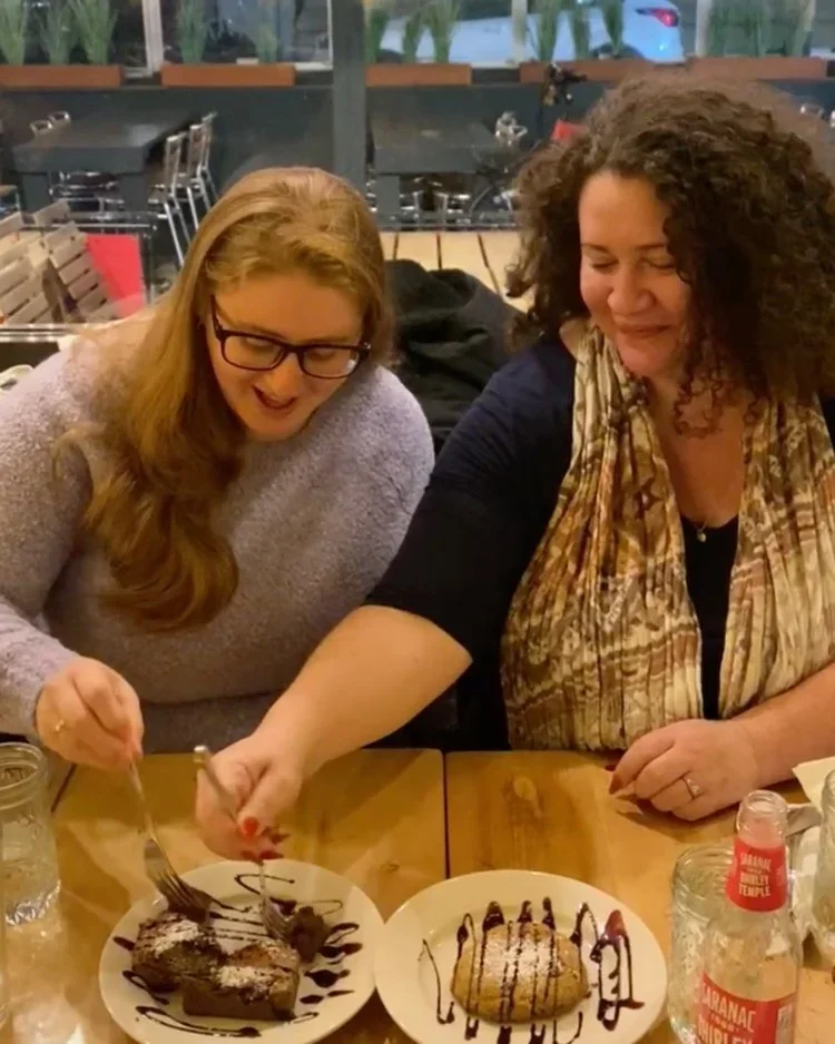 Two women sharing a chocolate dessert and smiling at a wooden table in a restaurant.