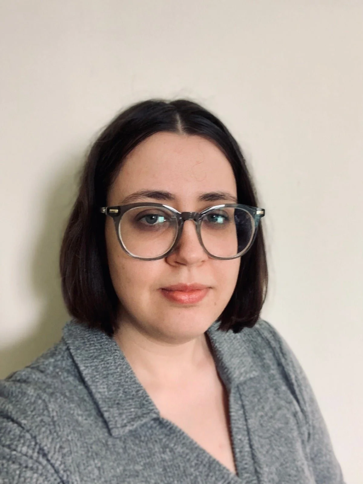 A woman with shoulder-length dark hair, wearing large round glasses, a gray blazer, and a gray top, standing against a plain off-white wall.