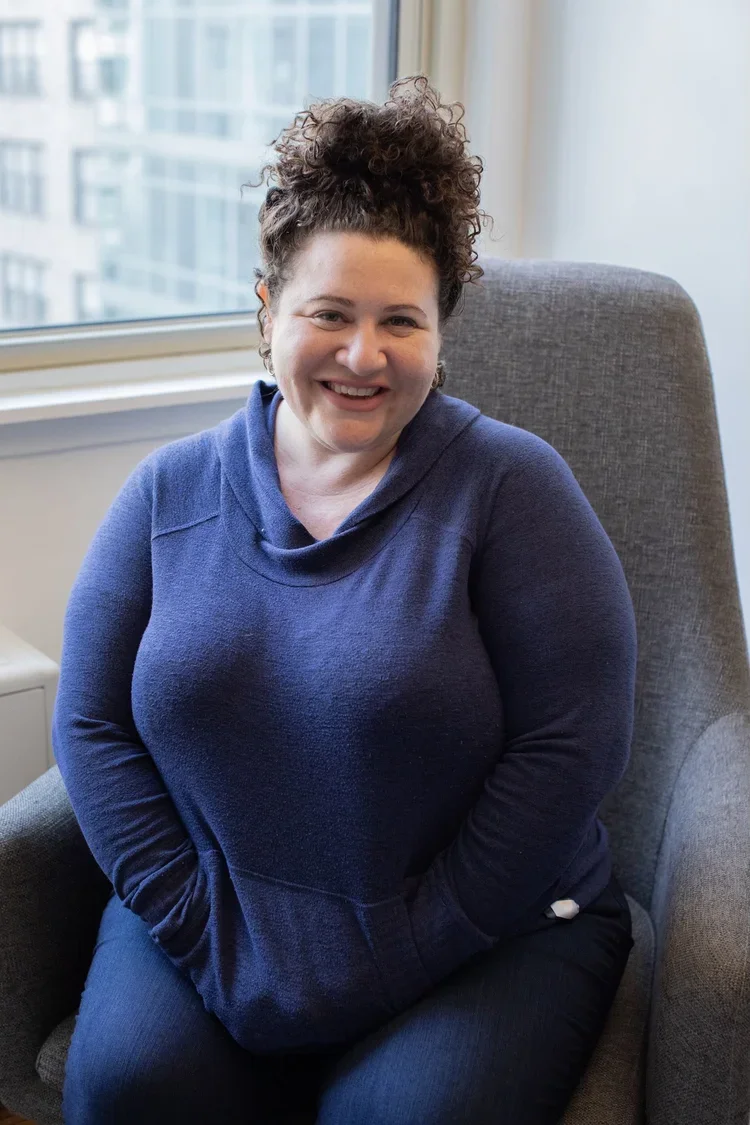 A woman with curly hair in a bun, wearing a blue hoodie, sitting in a gray armchair near a window with a city view, smiling at the camera.
