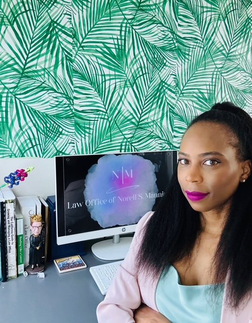 African American Woman with dark hair and pink lipstick sitting at a desk in front of a computer displaying a logo that reads "Law Office of Norell S. Manning" with a colorful background. Behind her is a wall with green tropical leaf wallpaper.