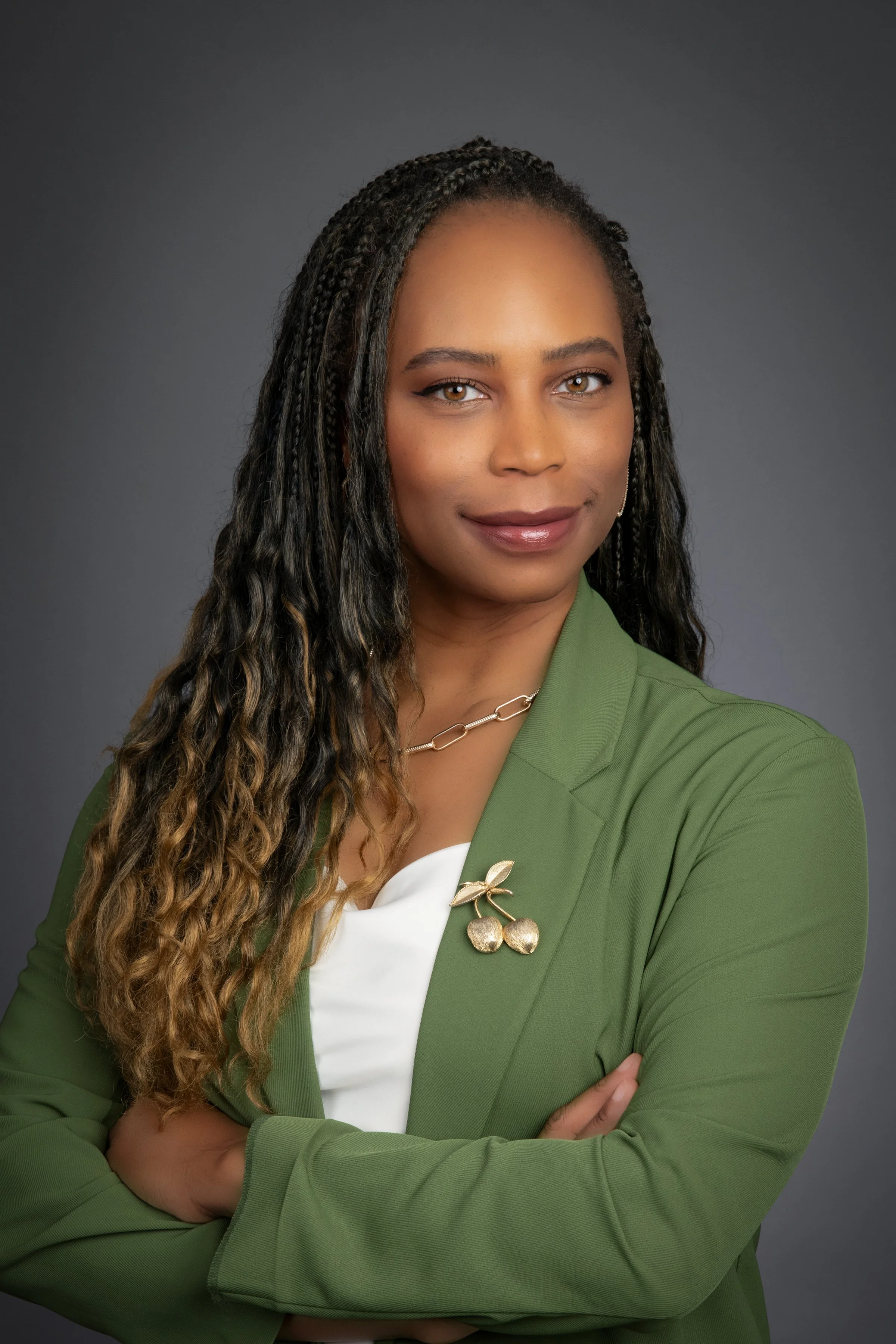A woman with long curly hair wearing a green blazer over a white top, with gold cherry-shaped brooch and necklace, standing against a gray background.