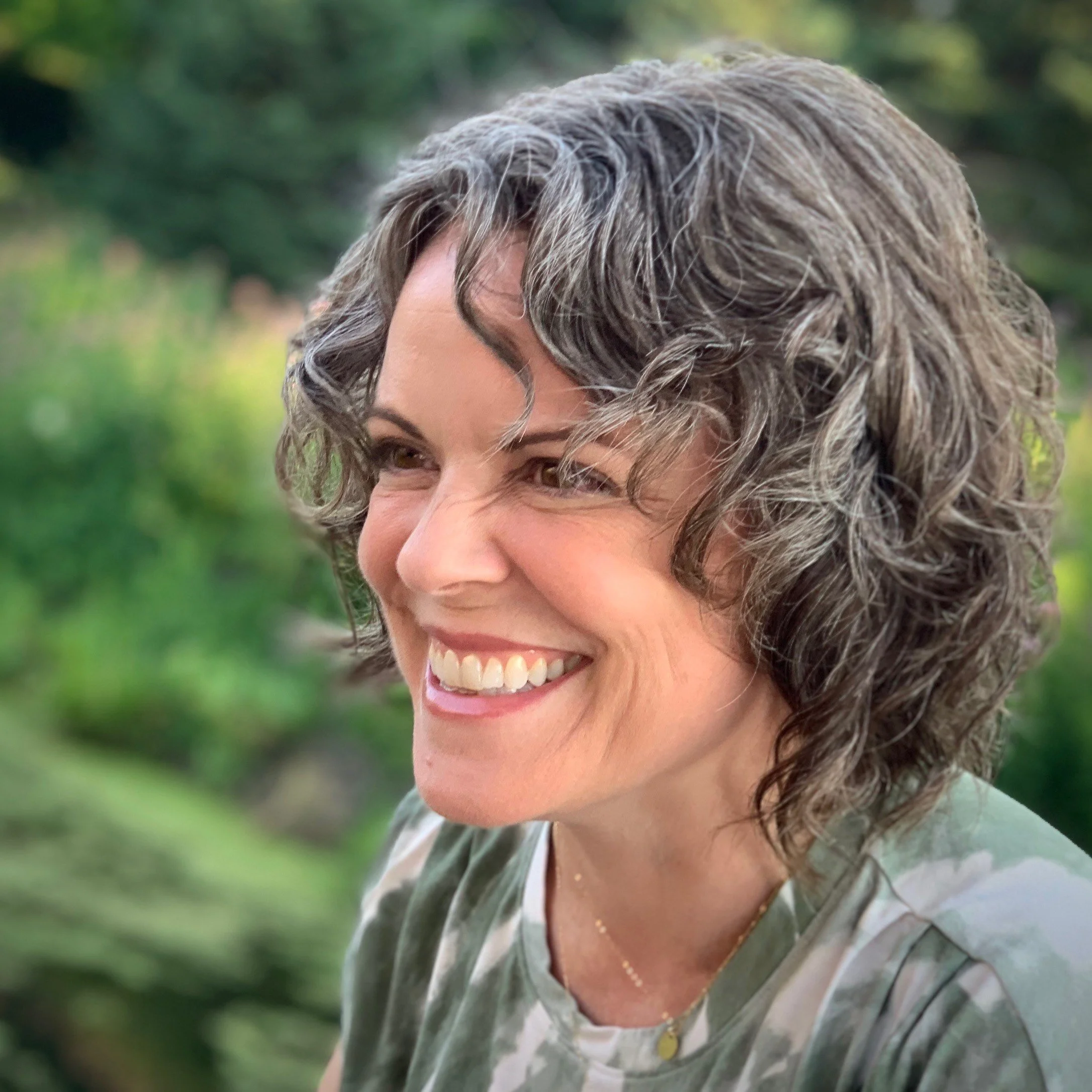 Close-up photograph of author Lauren Fischer, who has short, curly brown and gray hair; she is smiling outdoors with a green, blurred background.