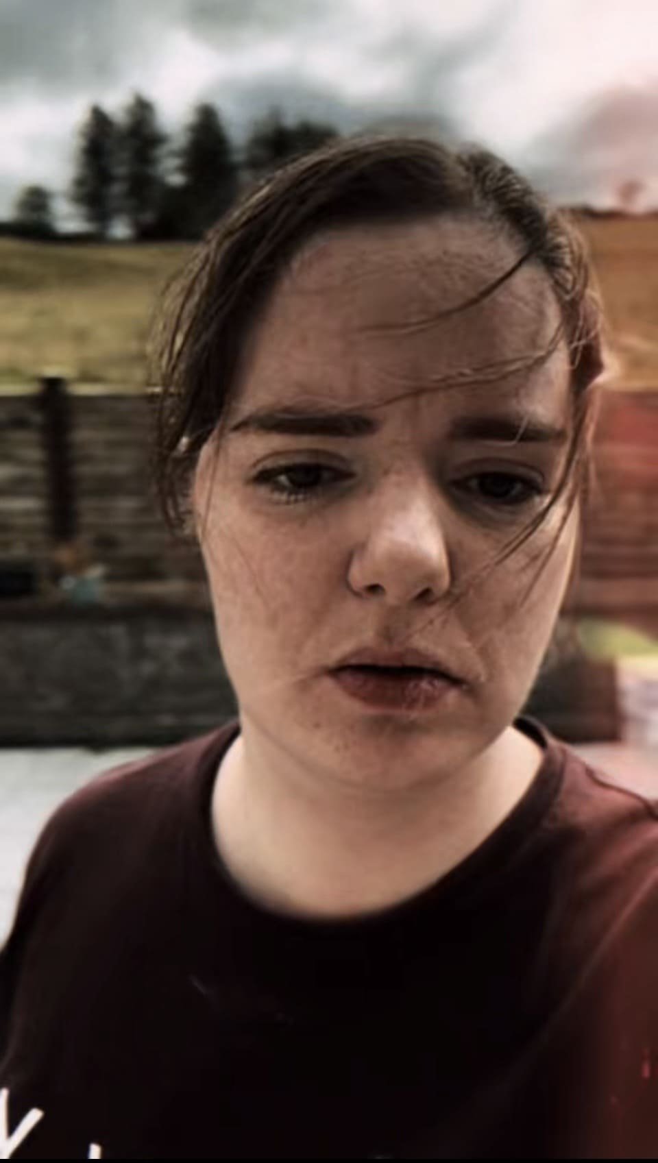 Close-up of a woman with a somber expression, brown hair, and wearing a dark shirt outdoors during overcast weather, with blurred background trees and wooden structures.