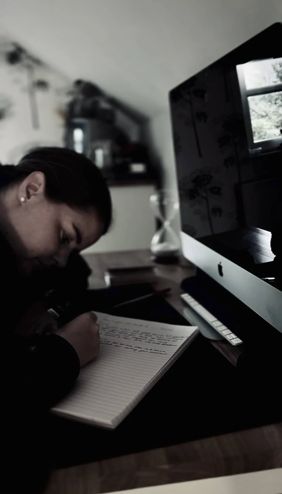 A woman writing in a notebook on a desk next to an Apple iMac computer in a dimly lit room.