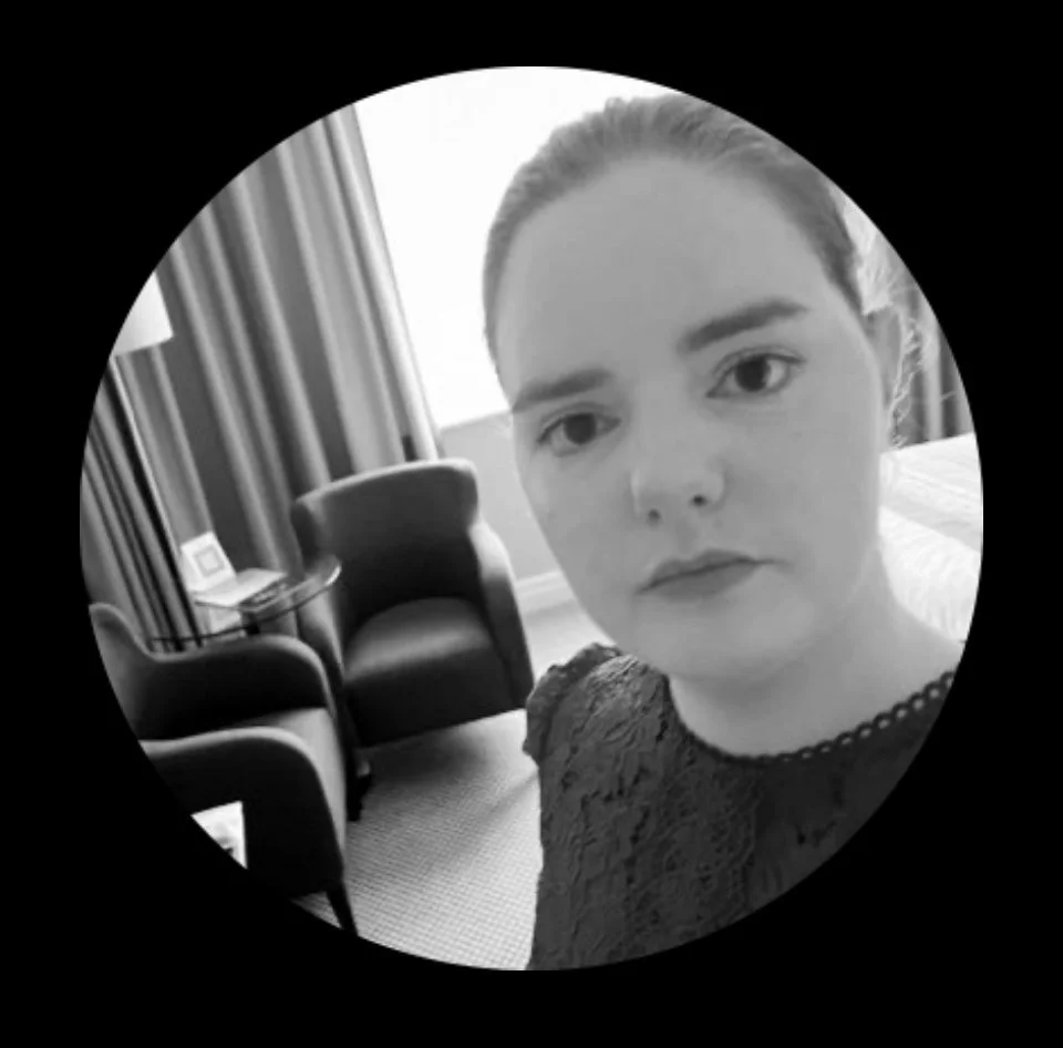 Black and white selfie of a woman with light skin, dark eyes, and hair tied back, wearing a lace dress, sitting in a hotel room with armchairs, curtains, and a window in the background.