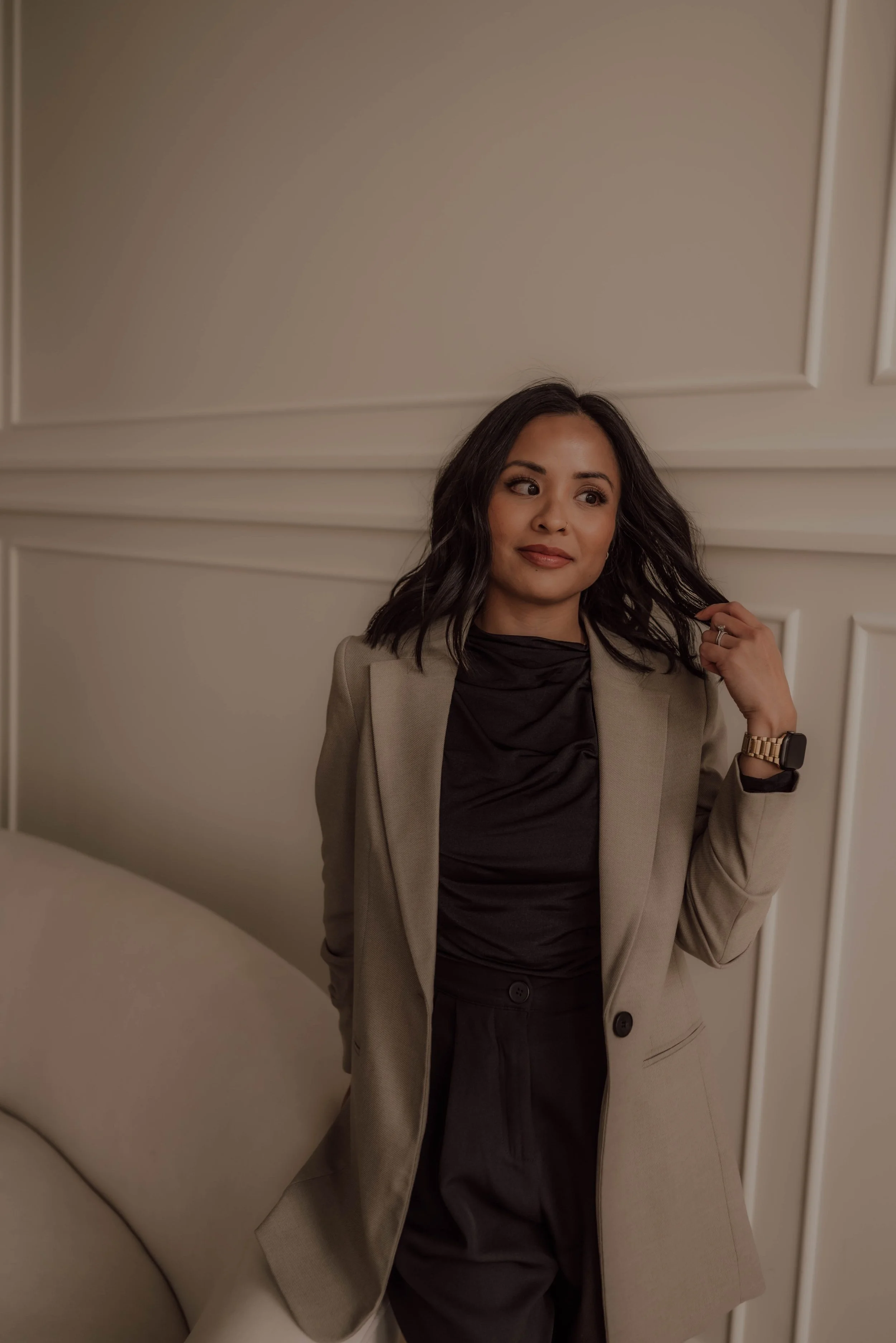 A woman with black hair wearing a beige blazer, black top, and black pants, standing by a cream-colored sofa, touching her hair with her right hand, in a room with white paneled walls.