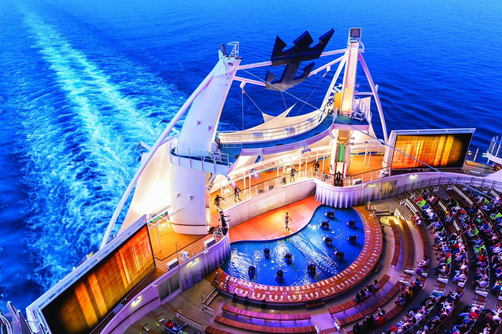 Aerial view of the deck on a cruise ship at sunset with a pool and seating area, and passengers enjoying the outdoor amenities.