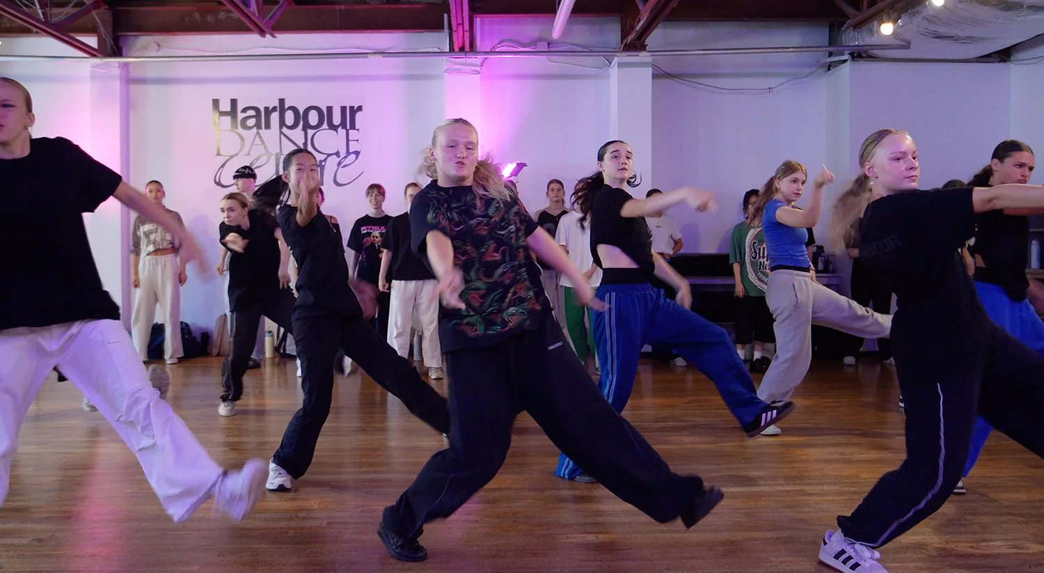 A group of young women practicing dance moves in a studio with wooden floors and purple lighting, some standing against the wall and others in the foreground actively dancing.