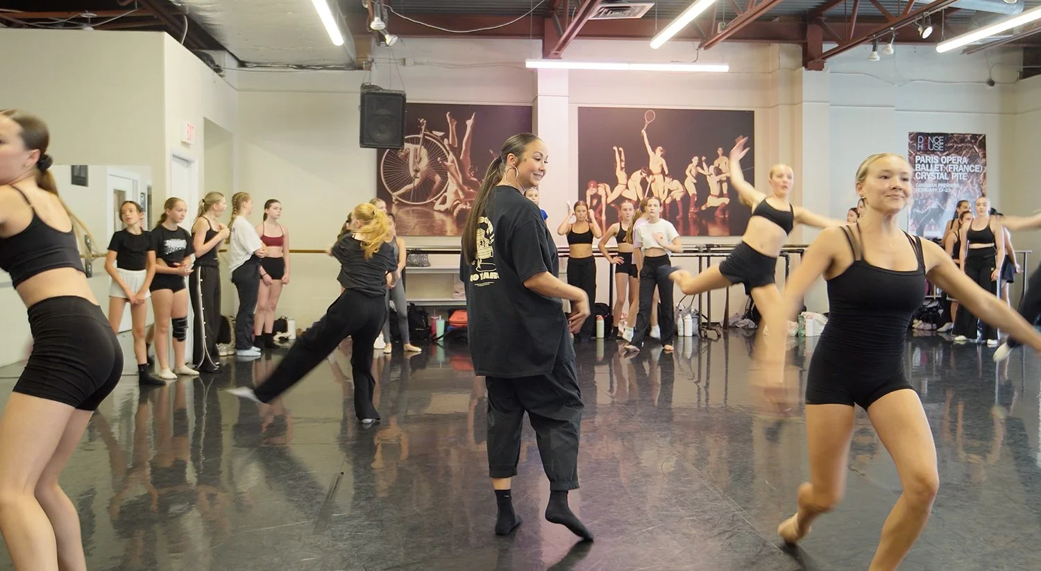 Young women participating in a dance class in a studio, some practicing and others observing, with walls decorated with dance posters.