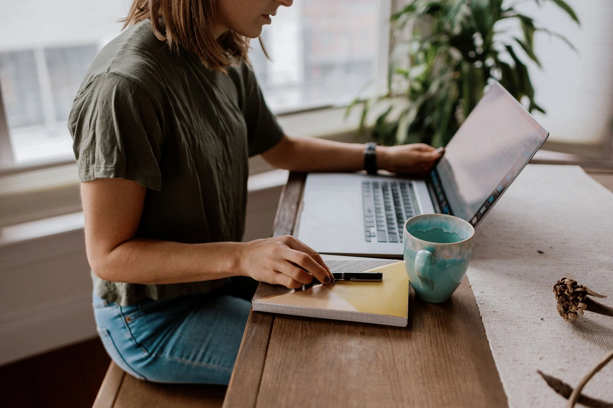 Woman working on a laptop at a wooden desk, with a notebook and pen nearby, along with a teal coffee mug.