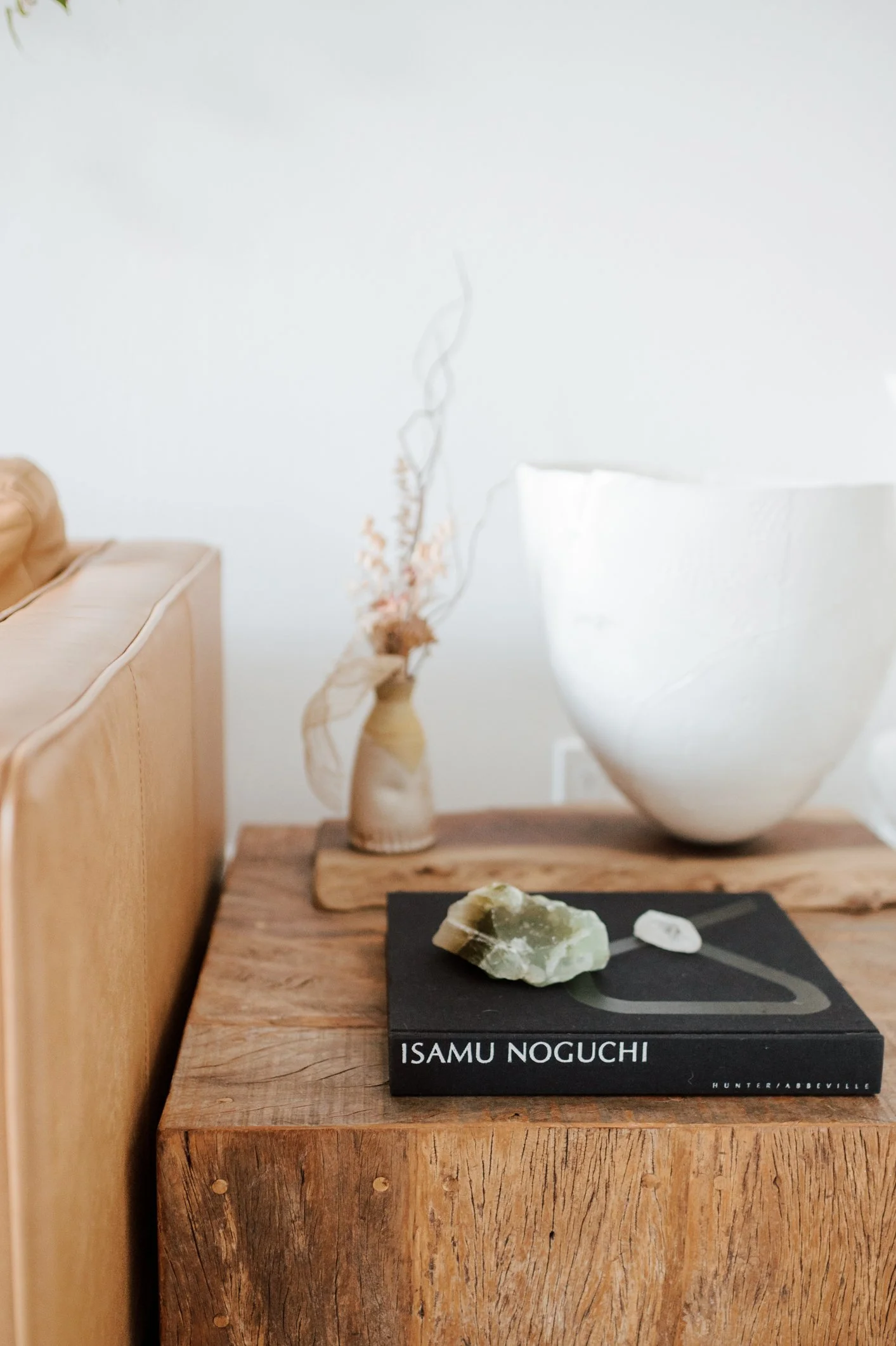 Wooden table with book titled 'Isamu Noguchi,' rock on book, tan leather couch, vase with dried flowers, and white sculptural bowl.