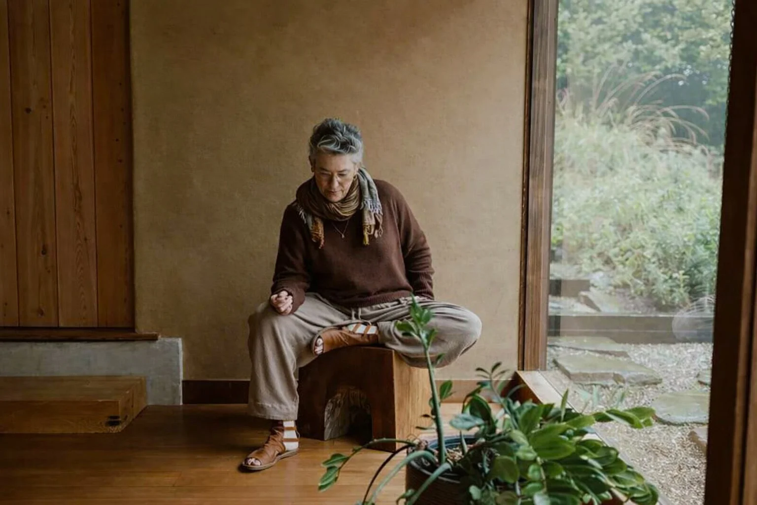 A woman with gray hair, wearing a brown sweater, beige pants, and brown sandals with socks, sitting cross-legged on a wooden block inside a cozy room with wooden paneling and a large window, looking down thoughtfully, with a green potted plant in the foreground and a garden visible outside.