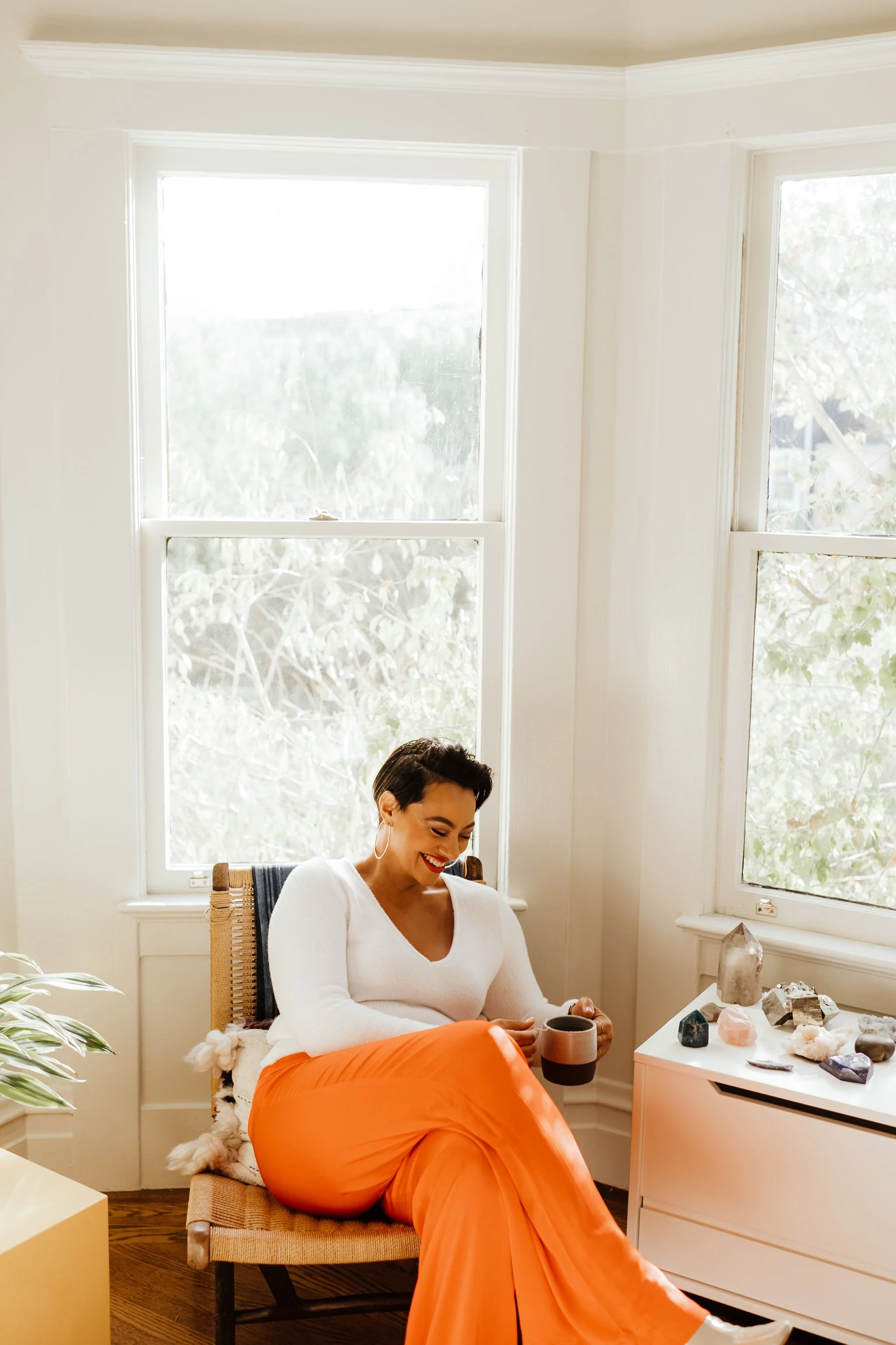 Midlife woman sitting in a cozy room, wearing a white top and orange pants, holding a mug, with crystals displayed on a small table nearby.