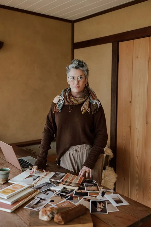 A woman with short gray hair, glasses, and a scarf standing behind a wooden table covered with photos, notebooks, and books in a cozy room with wooden walls.