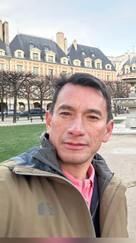 Hombre tomando una selfie en un parque en París, con edificios históricos y árboles sin hojas al fondo.