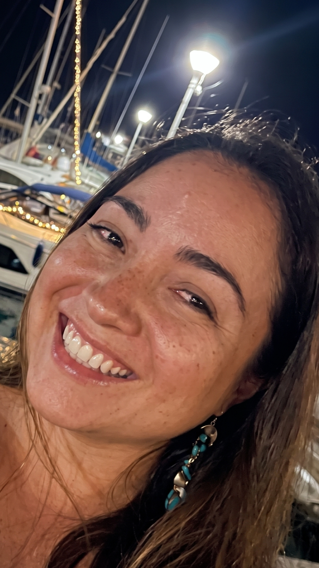 Retrato de una mujer sonriendo en un muelle con barcos, farolas y cielo nocturno, pelo castaño oscuro y accesorios de collar y aretes azules.