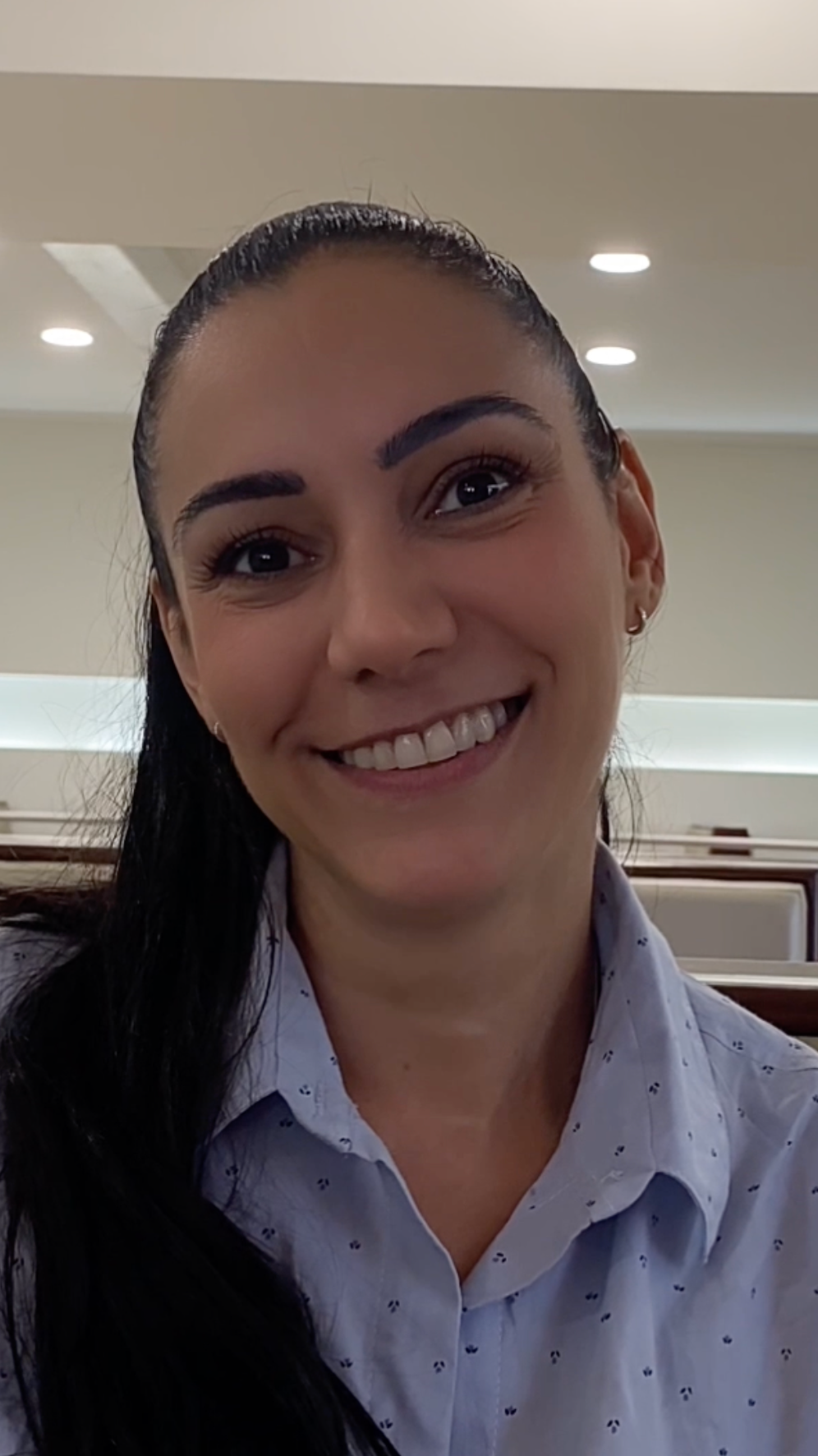 Retrato de una joven mujer sonriendo, con cabello oscuro y coleta, usando blusa de botones en un lugar cerrado con luces en el techo.