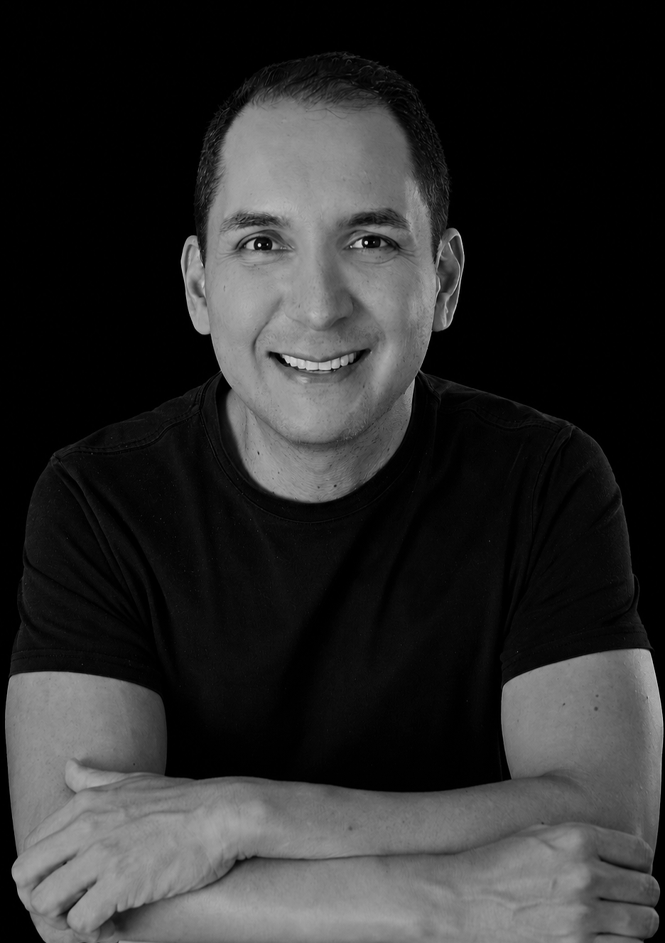 Retrato en blanco y negro de un hombre sonriendo, con cabello corto y camisa negra, con fondo negro.