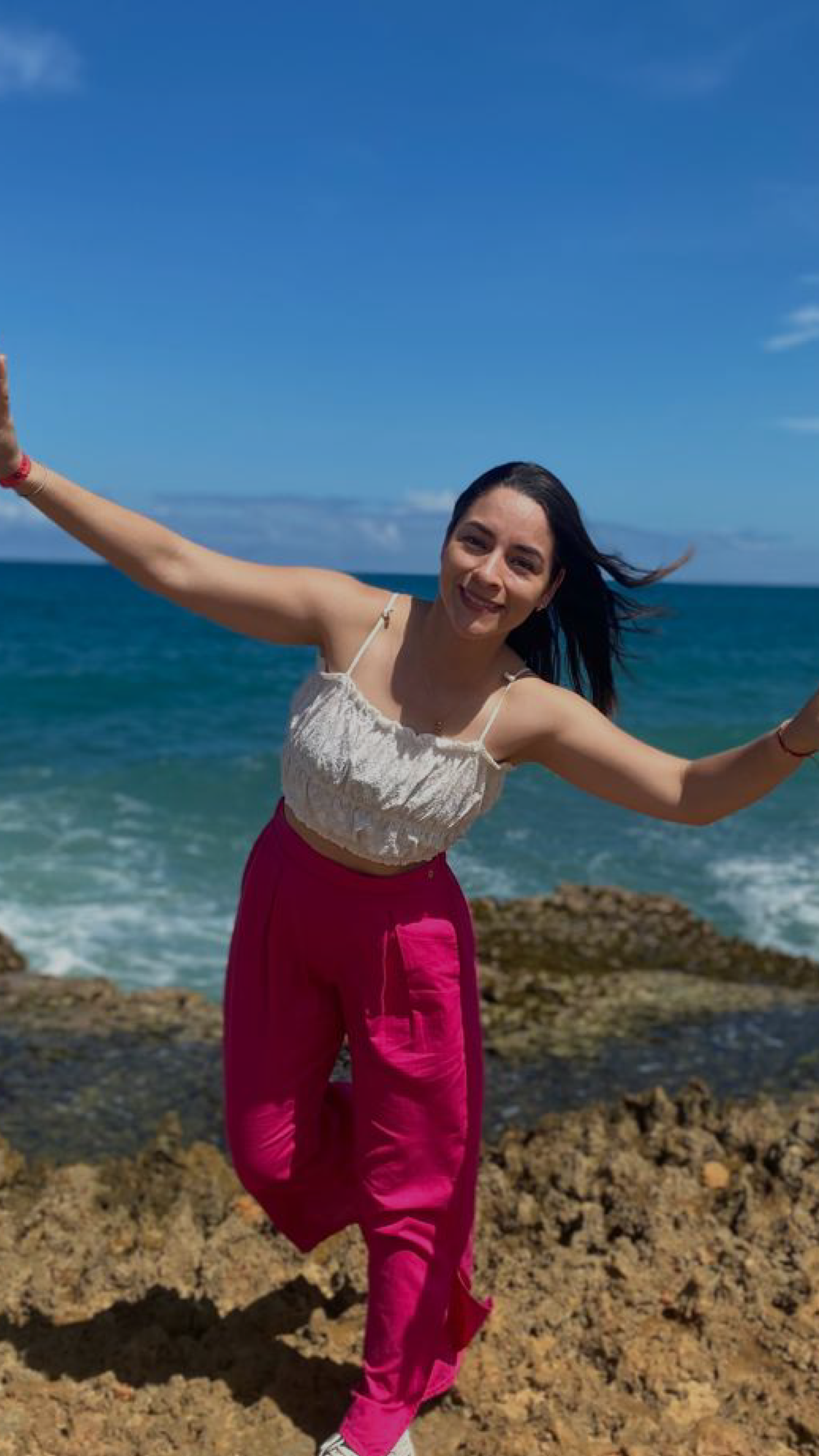 Una joven sonriente de piel clara, con cabello negro y largo, viste una blusa blanca sin mangas y pantalones deportivos rosados, posando en una playa con rocas y el océano al fondo bajo un cielo despejado.
