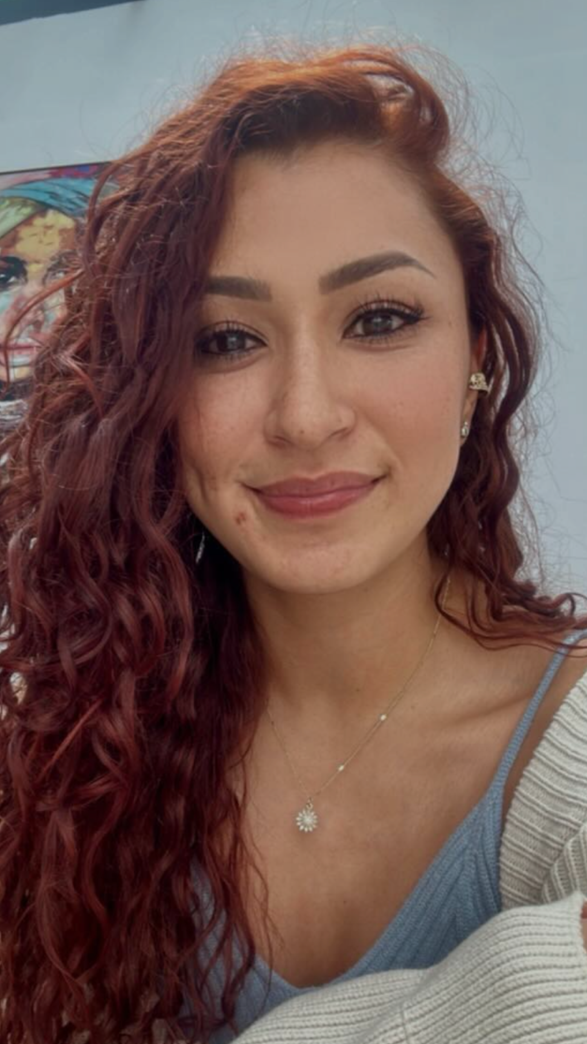 Retrato de una mujer con cabello rizado de color rojo y piel clara, sonriendo y usando una blusa azul y un collar. Fondo neutro con una pintura en la pared.