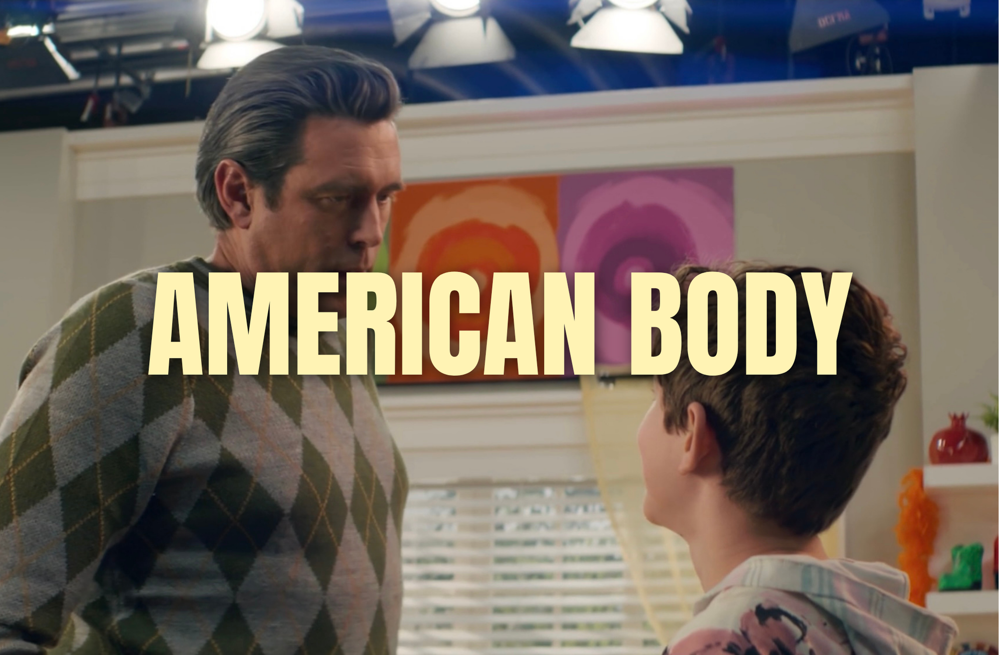 Scene from the TV show 'American Body' featuring two characters, a man with dark hair in a colorful argyle sweater and a boy with short hair in a patterned shirt, in a brightly decorated room with colorful artwork on the wall.
