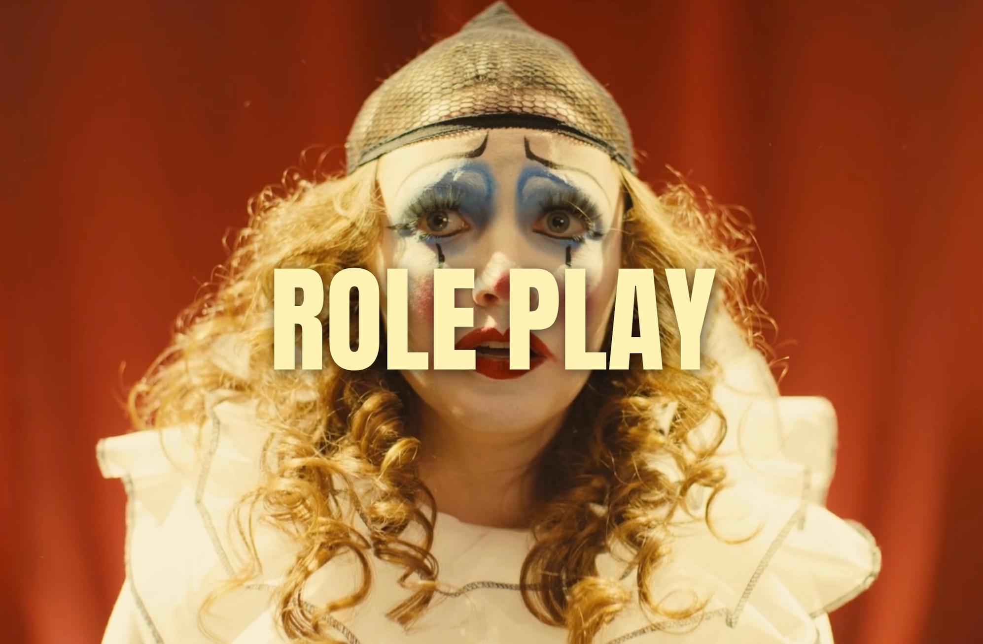 A person dressed as a clown with curly red hair, white face makeup with blue eyeshadow, red lips, and drawn tears, wearing a striped clown hat and a ruffled white costume. The background is red, and the words "ROLE PLAY" are overlaid on the image.
