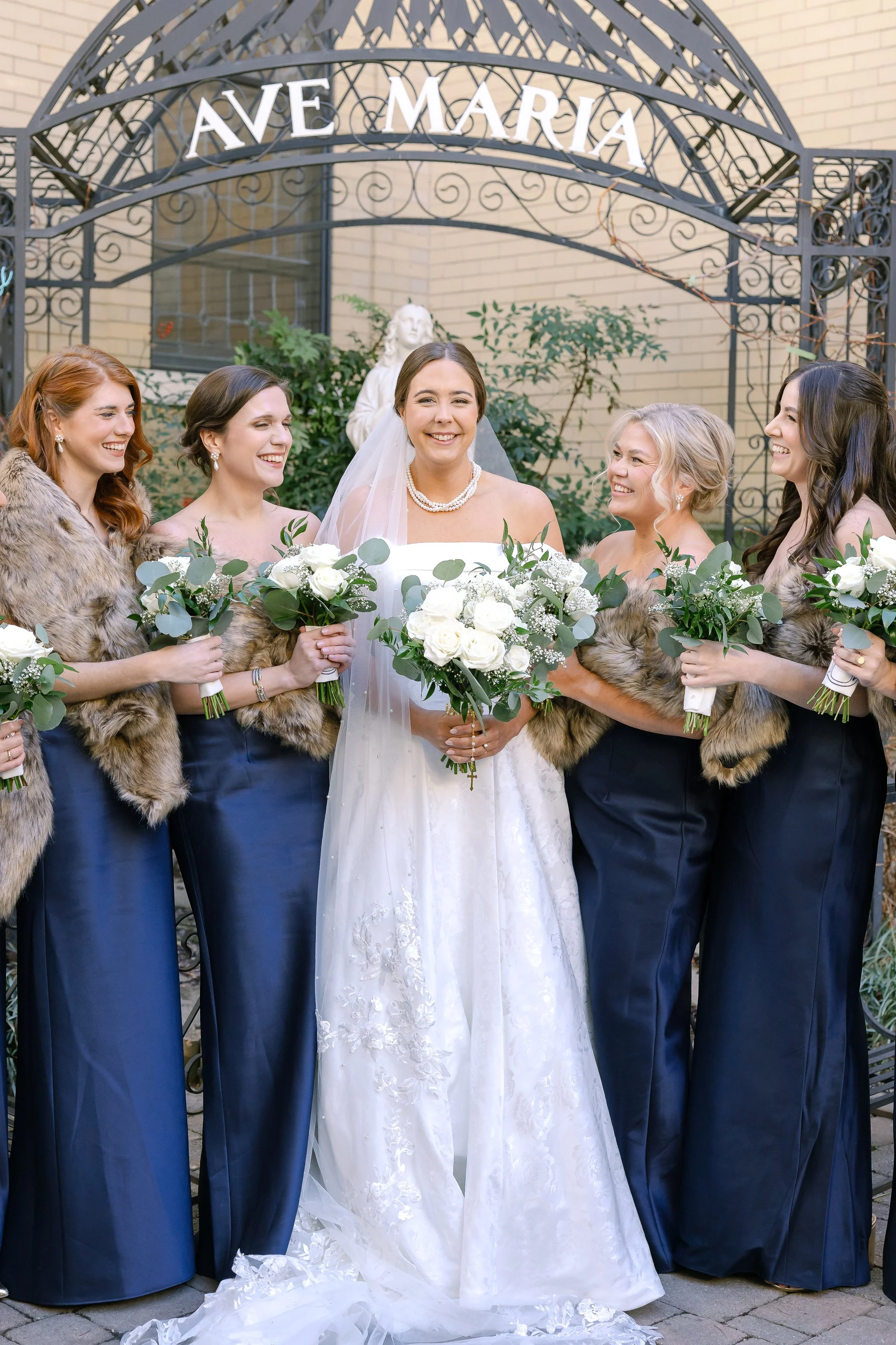 Wedding celebration outside with a bride and groom kissing, surrounded by cheerful friends, some holding bouquets, in front of a brick building.