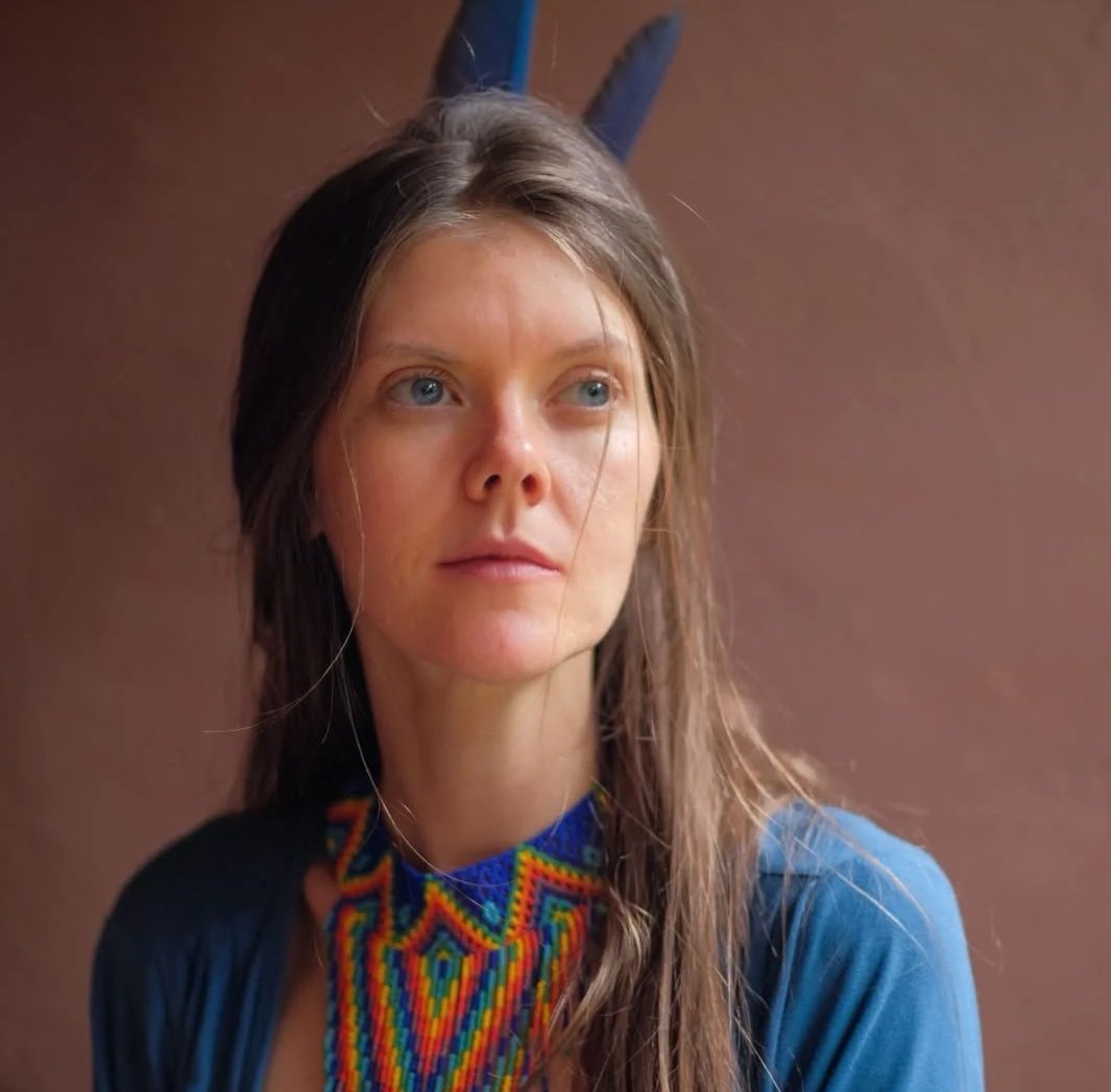 Cranial Bliss Therapy client, young woman with long brown hair and blue eyes gazing to the side, wearing a colourful beaded necklace and a blue top, standing against a plain brown background.