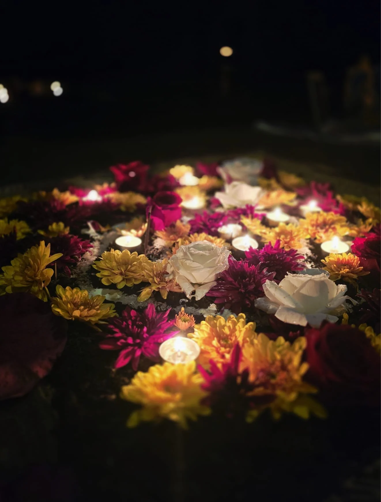 Nighttime scene of a floral arrangement with candles, featuring white roses, pink, yellow, and purple flowers, with candles illuminating the flowers.