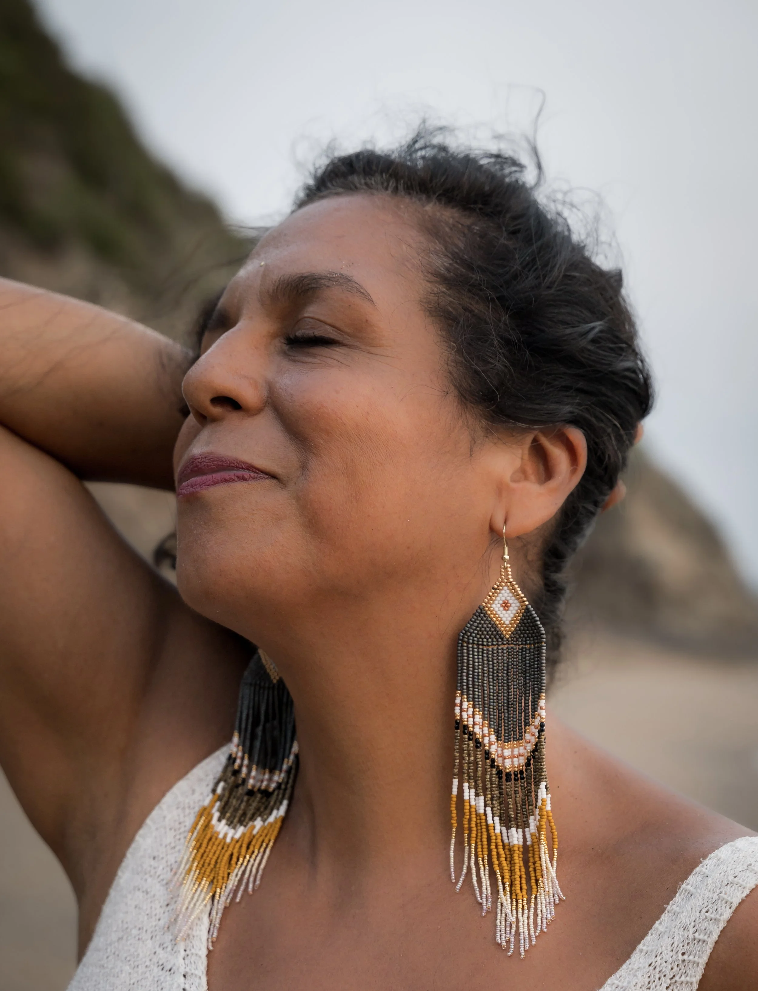 Close-up of a woman with short black hair, wearing beaded earrings and a beaded necklace, with her eyes closed and her hand behind her head, outdoors.